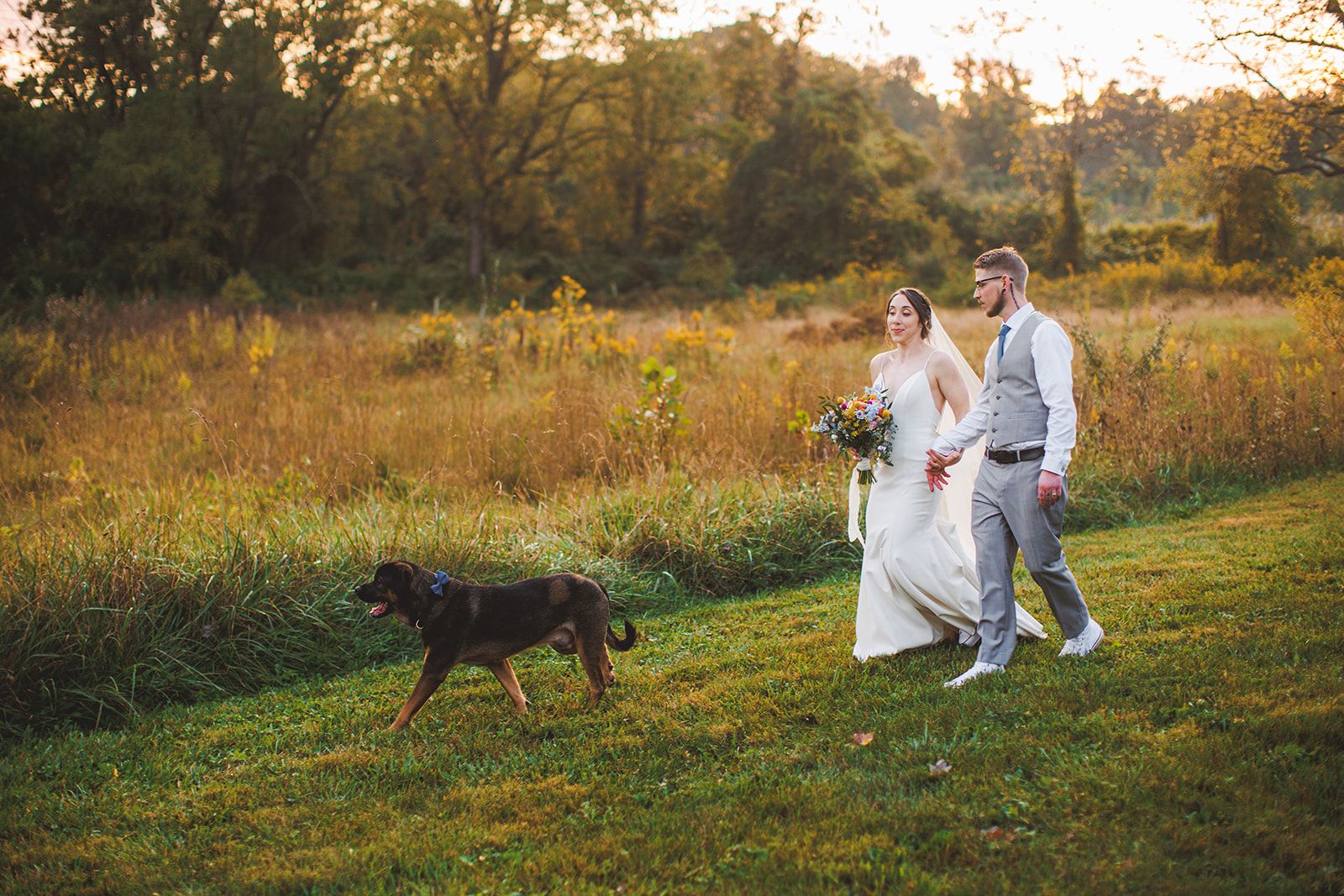 A dog trots ahead along the mown path through a golden field while the couple follows hand in hand.