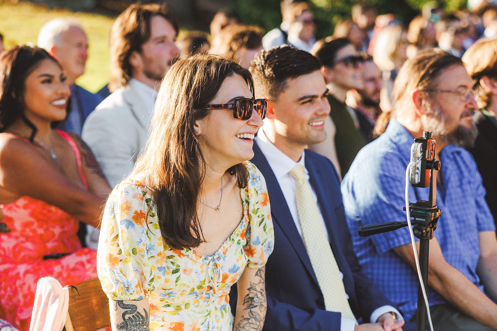 A woman in a floral dress and sunglasses laughs in her seat while the young man beside her in a navy suit smiles toward the front, low sun warming the rows of guests behind them.