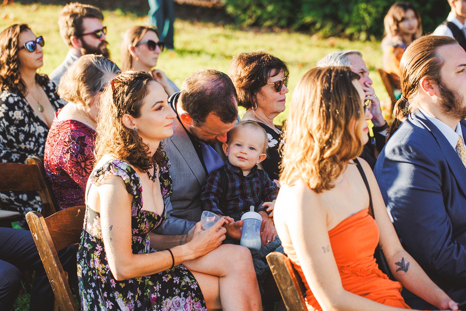 A man bends down to kiss a toddler on the head while the child, sippy cup in hand, looks straight into the camera from a row of seated guests.