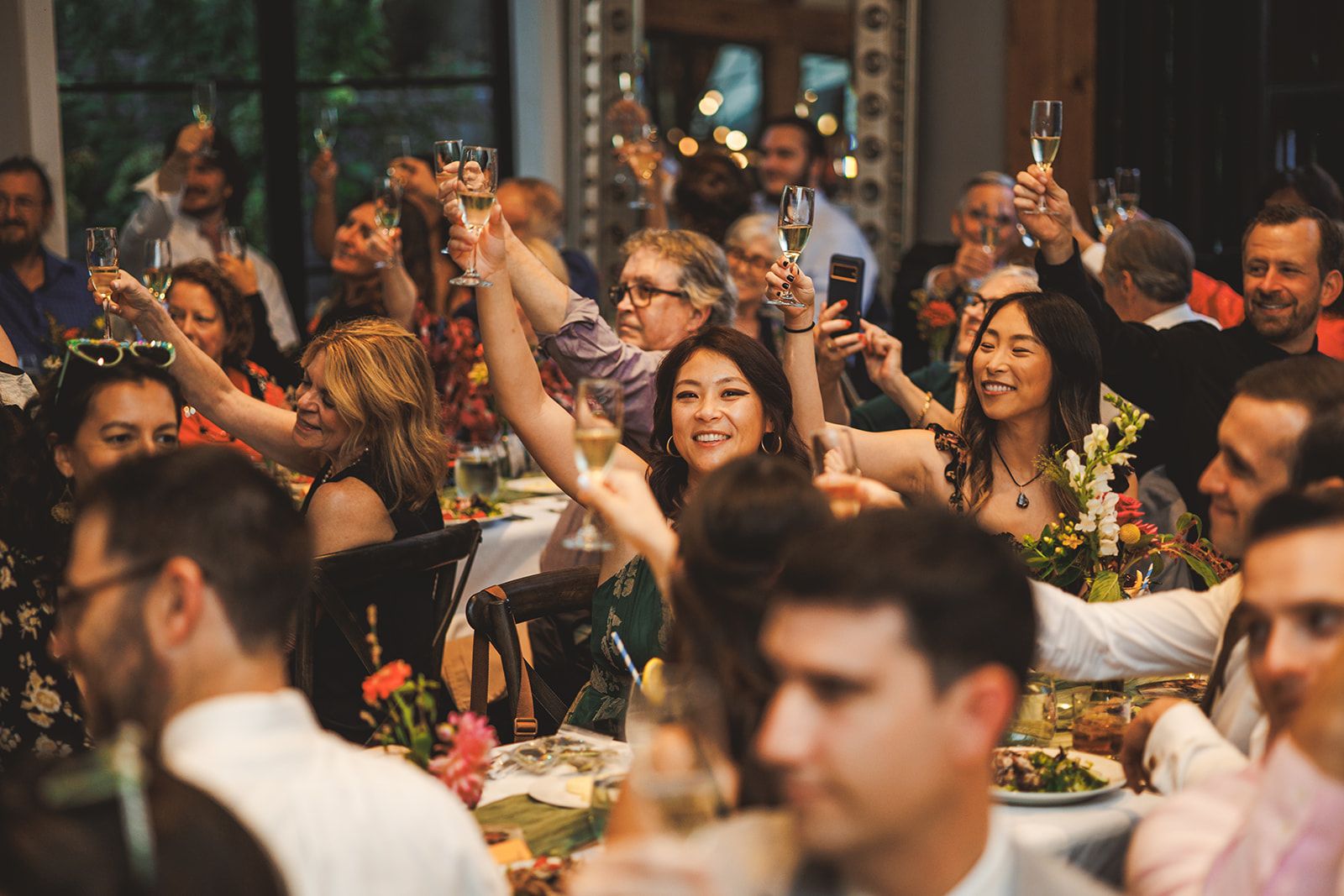 Guests around the reception tables raise champagne glasses in a toast, faces bright with laughter, one of them filming on a phone.