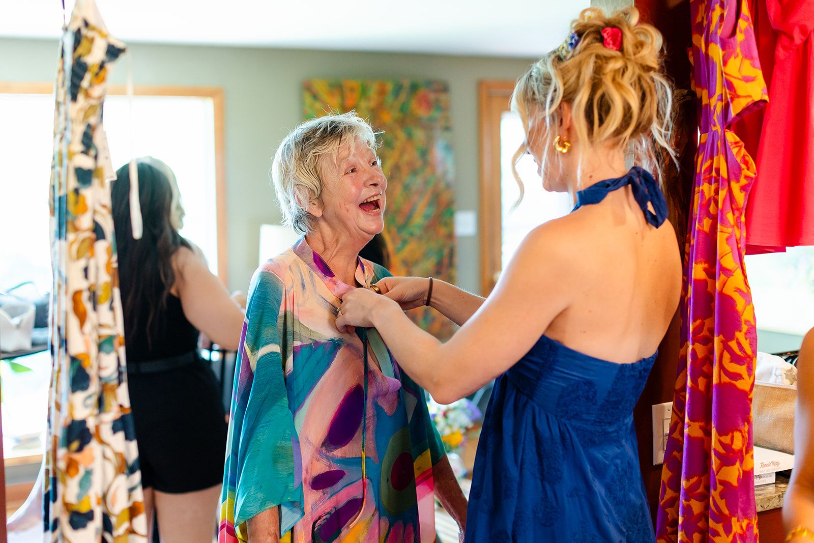 An older woman laughs with her chin raised while a younger woman in a navy dress fastens the colorful wrap at her neckline.
