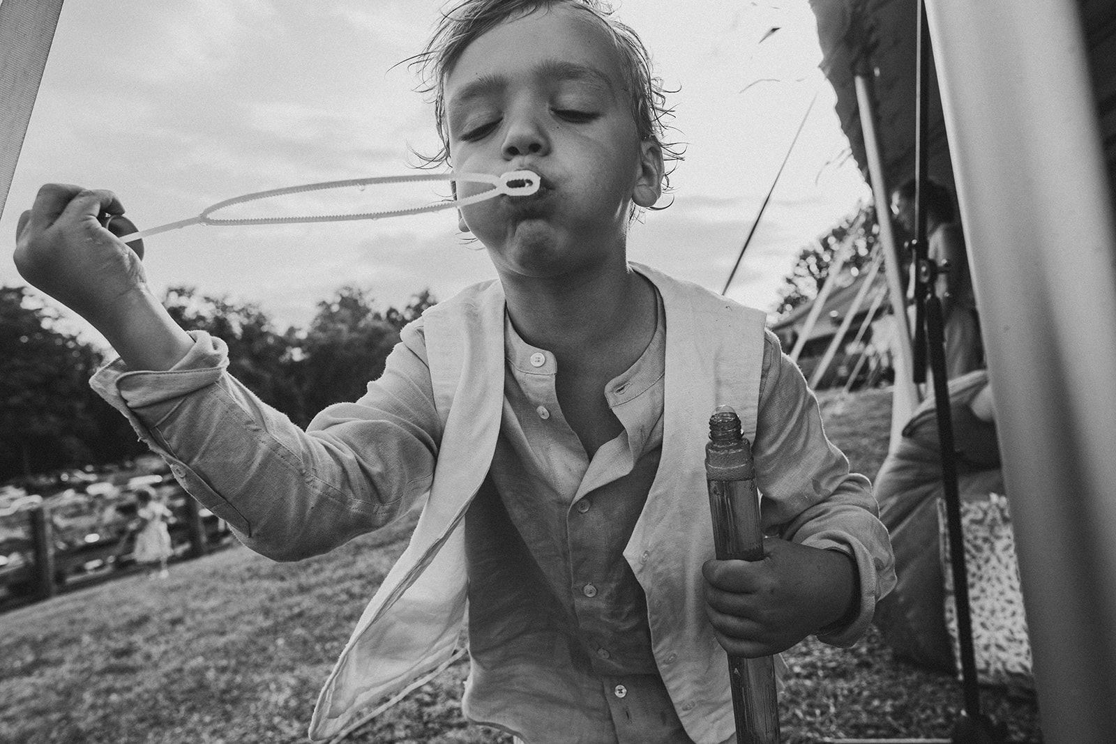 A child in an open vest blows through a bubble wand with the bubble bottle still gripped in the other hand.