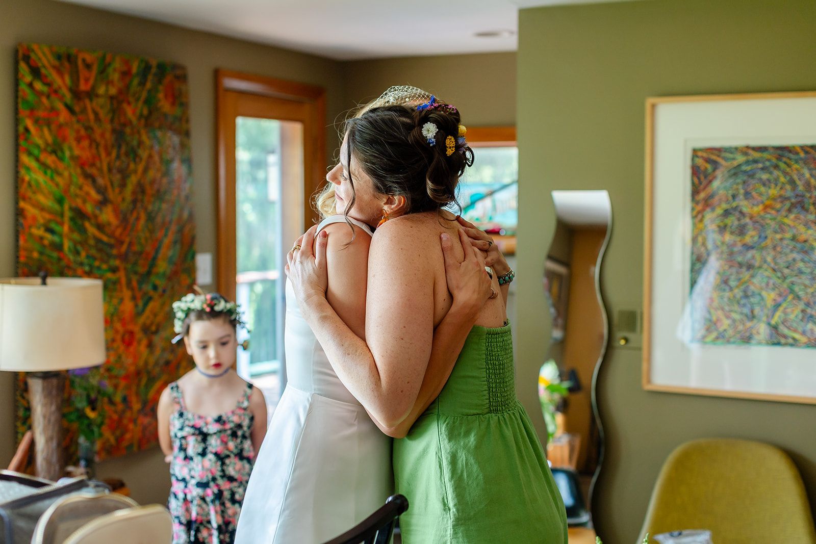 A bride in a white gown hugs a woman in a green dress while a young girl in a flower crown watches from behind.
