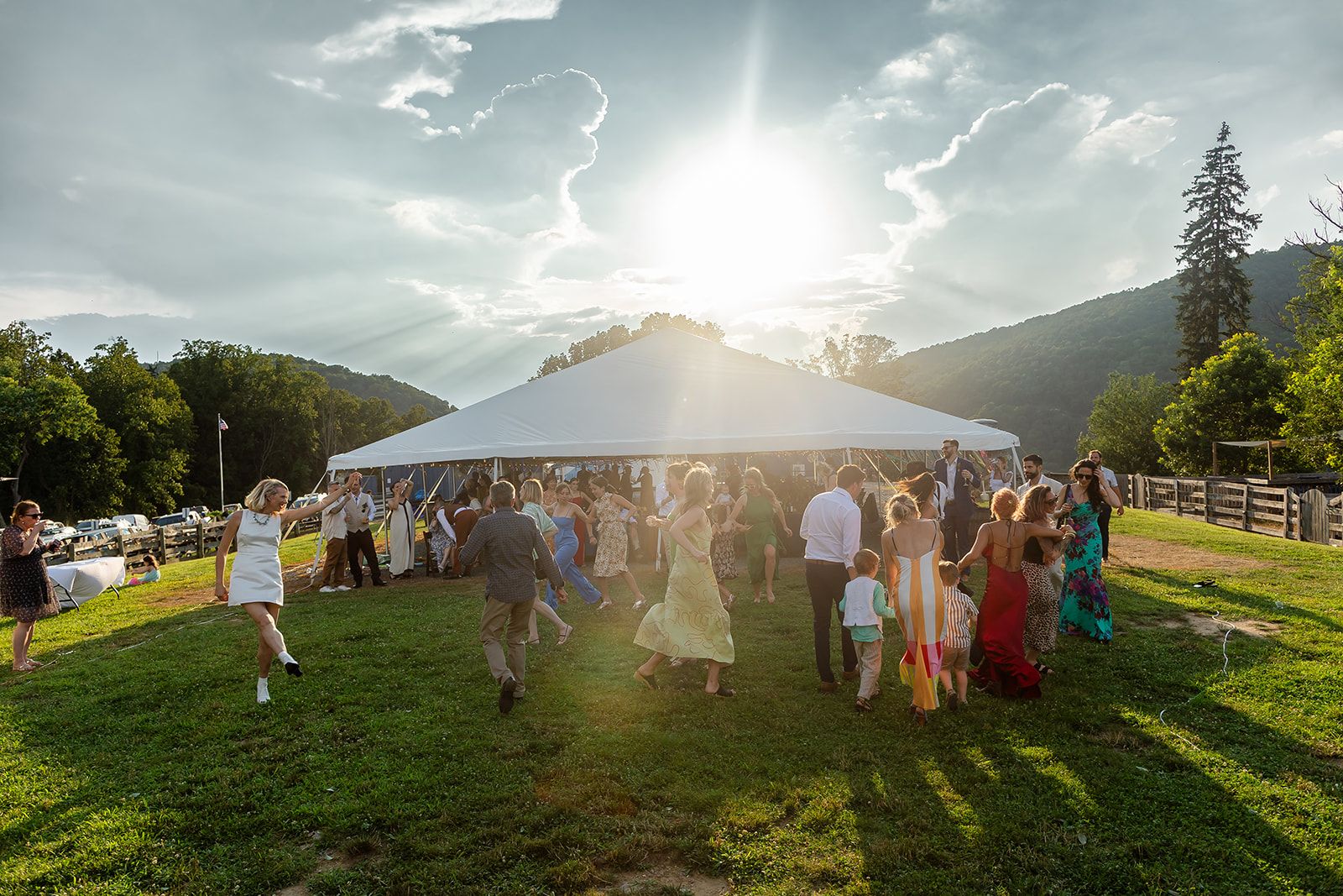 Guests stream across the grass toward an open-sided tent as the sun flares through the clouds behind it.