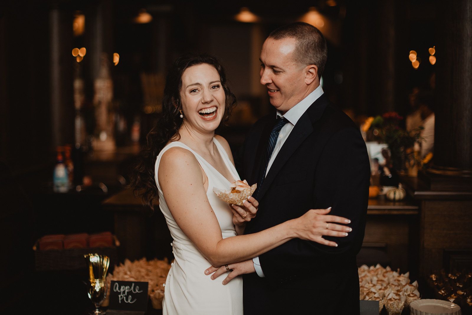 A bride laughs while holding a small pastry at the dessert table as the groom smiles down at her with his arm around her.