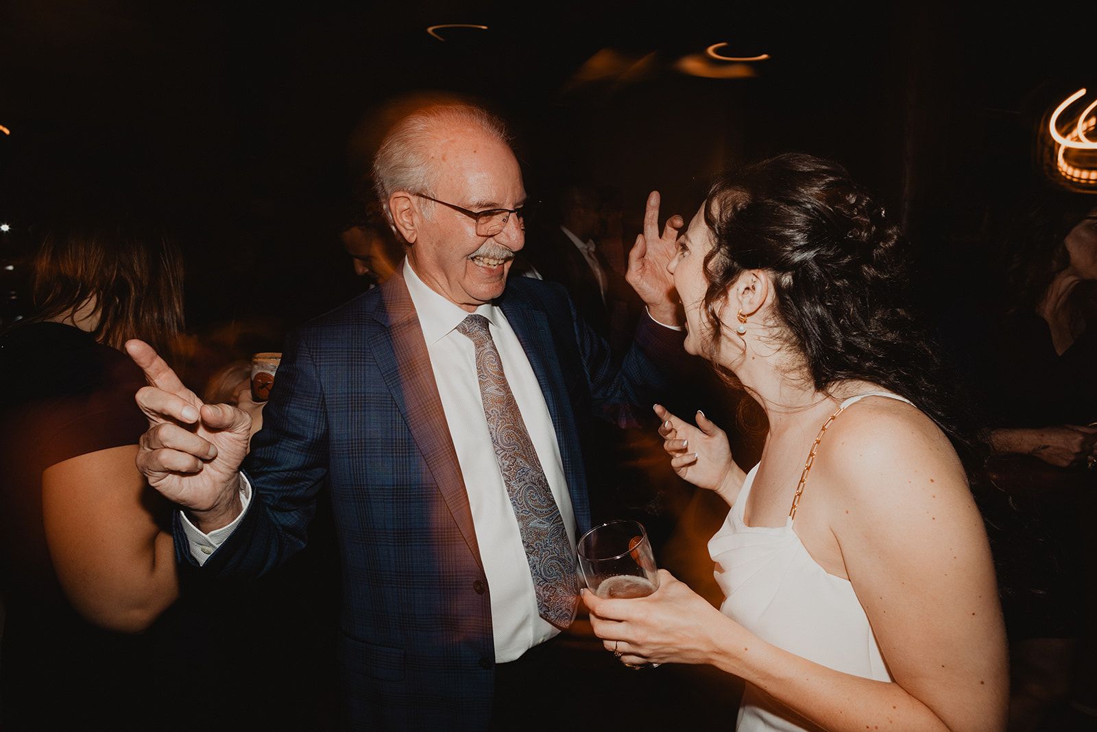 An older man in a plaid suit grins and points a finger in the air on the dance floor beside a laughing woman in a white dress holding a drink.