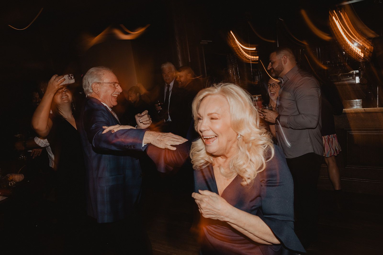 An older woman with blonde curls laughs mid-turn on the dance floor as an older man in a plaid suit reaches an arm out toward her.