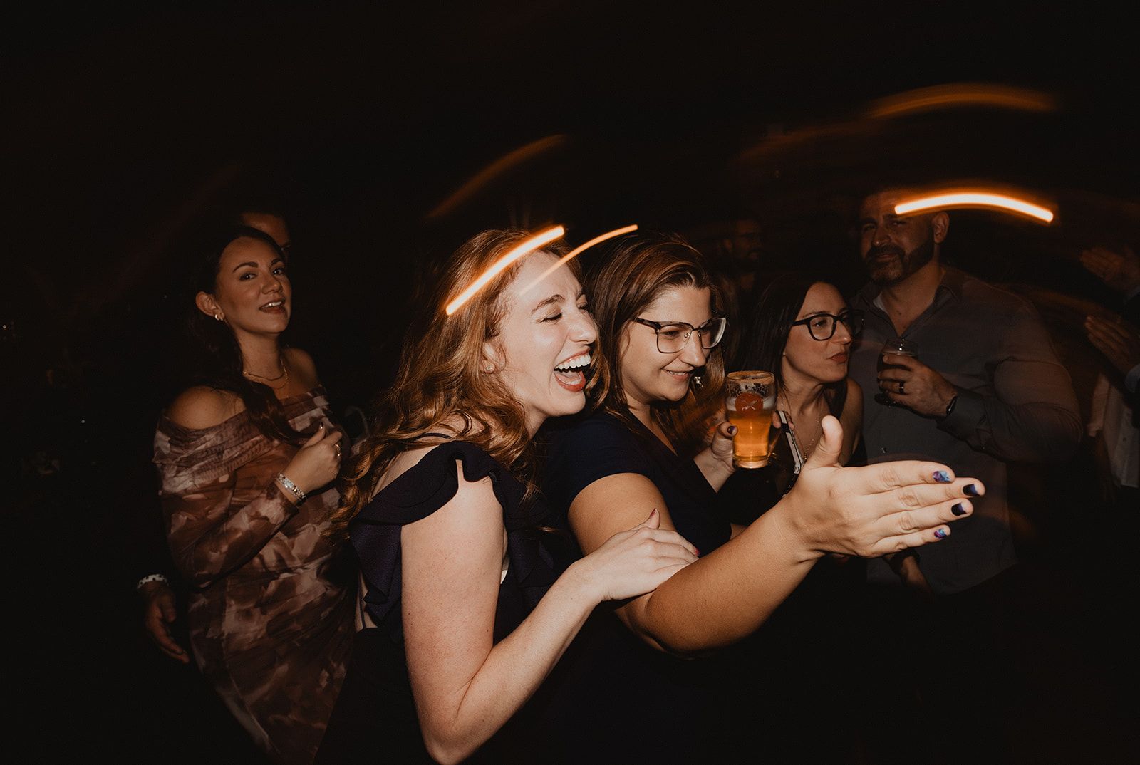 Two women laugh shoulder to shoulder at the reception, one reaching an open hand toward the camera and the other holding a beer, with more guests behind them.
