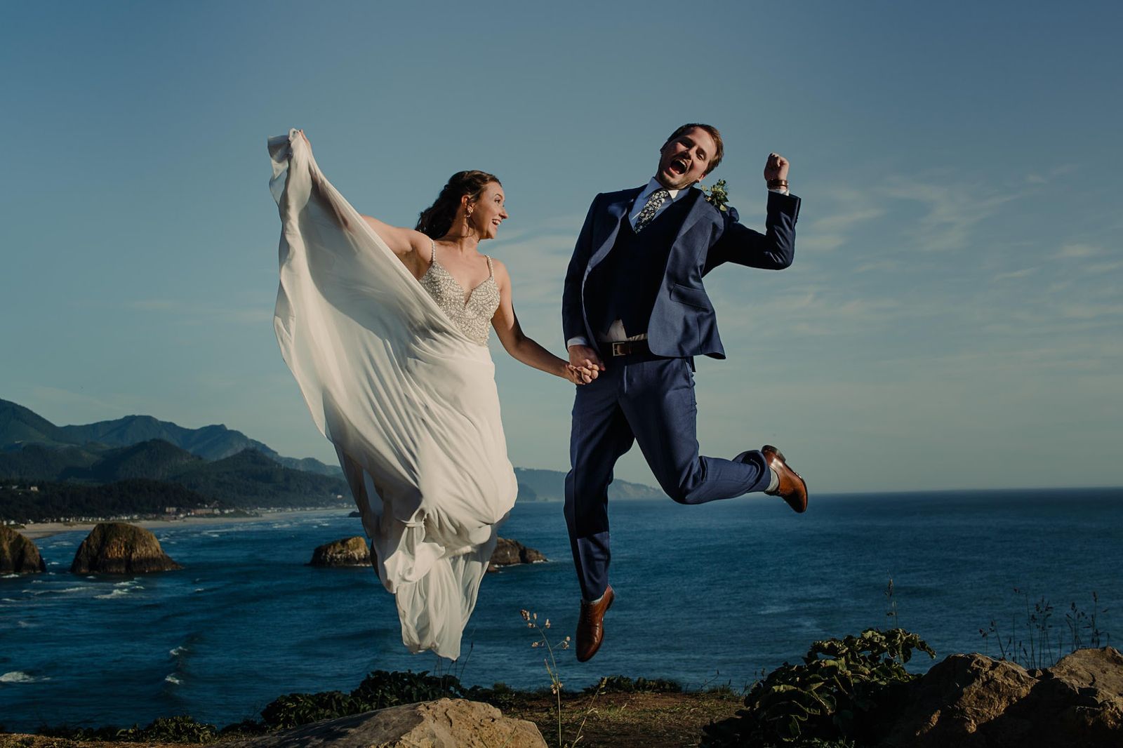 A couple in wedding clothes jump into the air hand in hand on a rocky bluff, both mouths open in a laugh, ocean and sea stacks behind them.