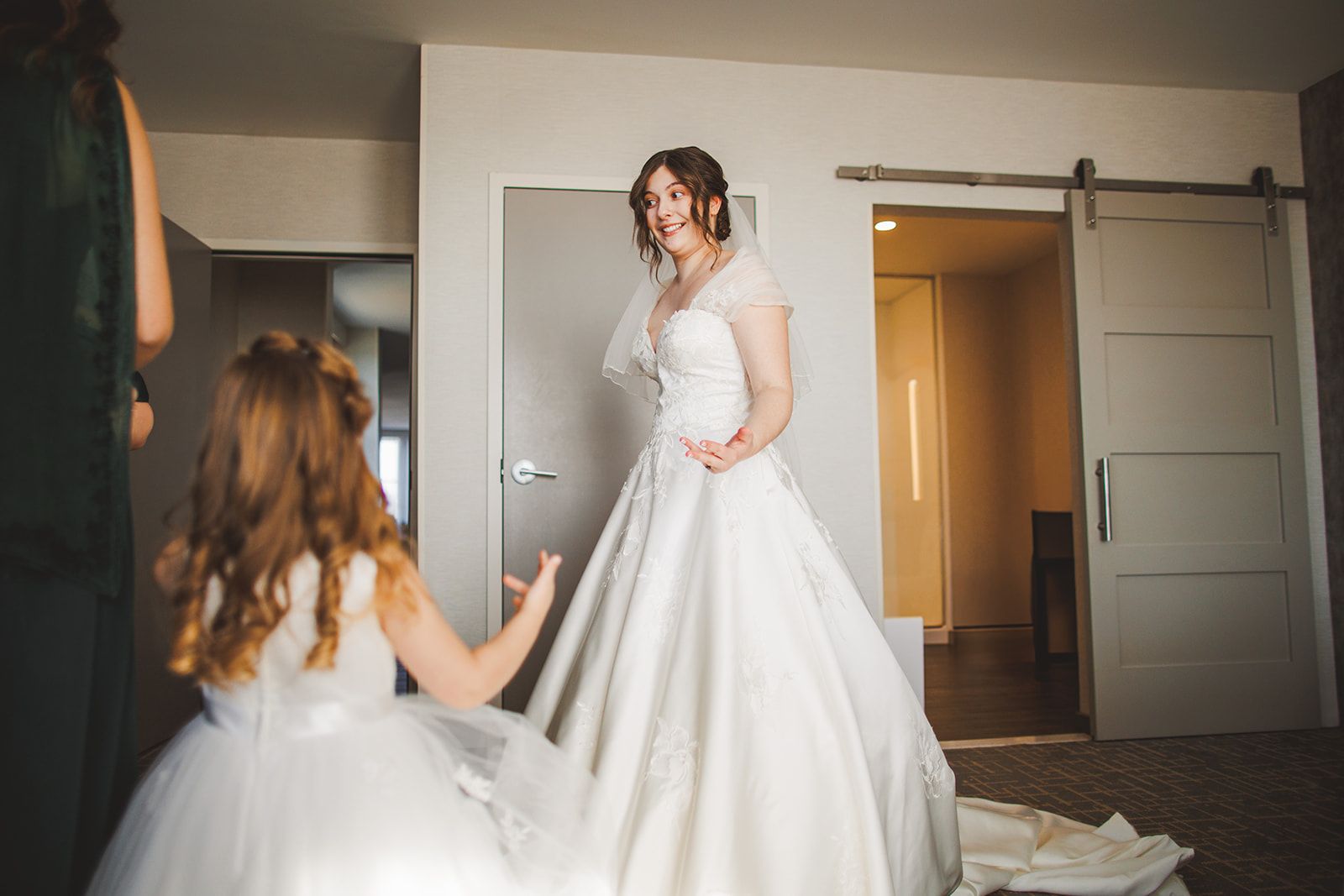 A young girl in a white dress reaches an open hand toward the bride's skirt while the bride turns and smiles at her.