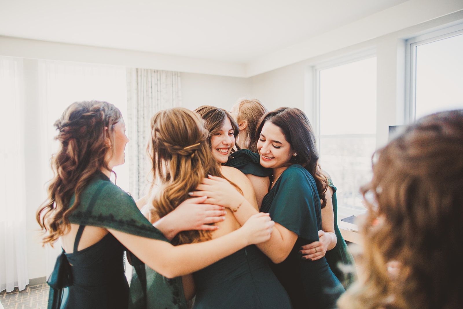Women in matching dark green dresses wrap each other in a group hug, one of them smiling with her eyes closed.
