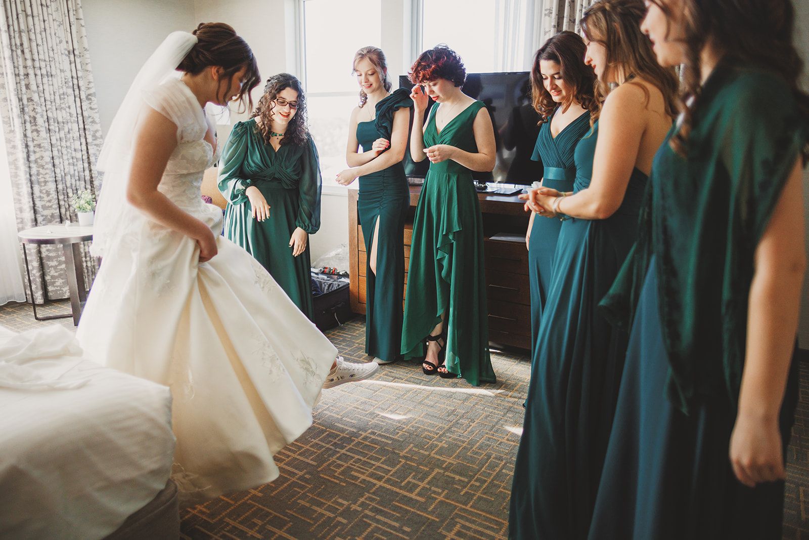 The bride bows her head and lifts a fold of her full skirt in a bright hotel room while bridesmaids in green stand around her, one grinning and one holding a hand to her brow.