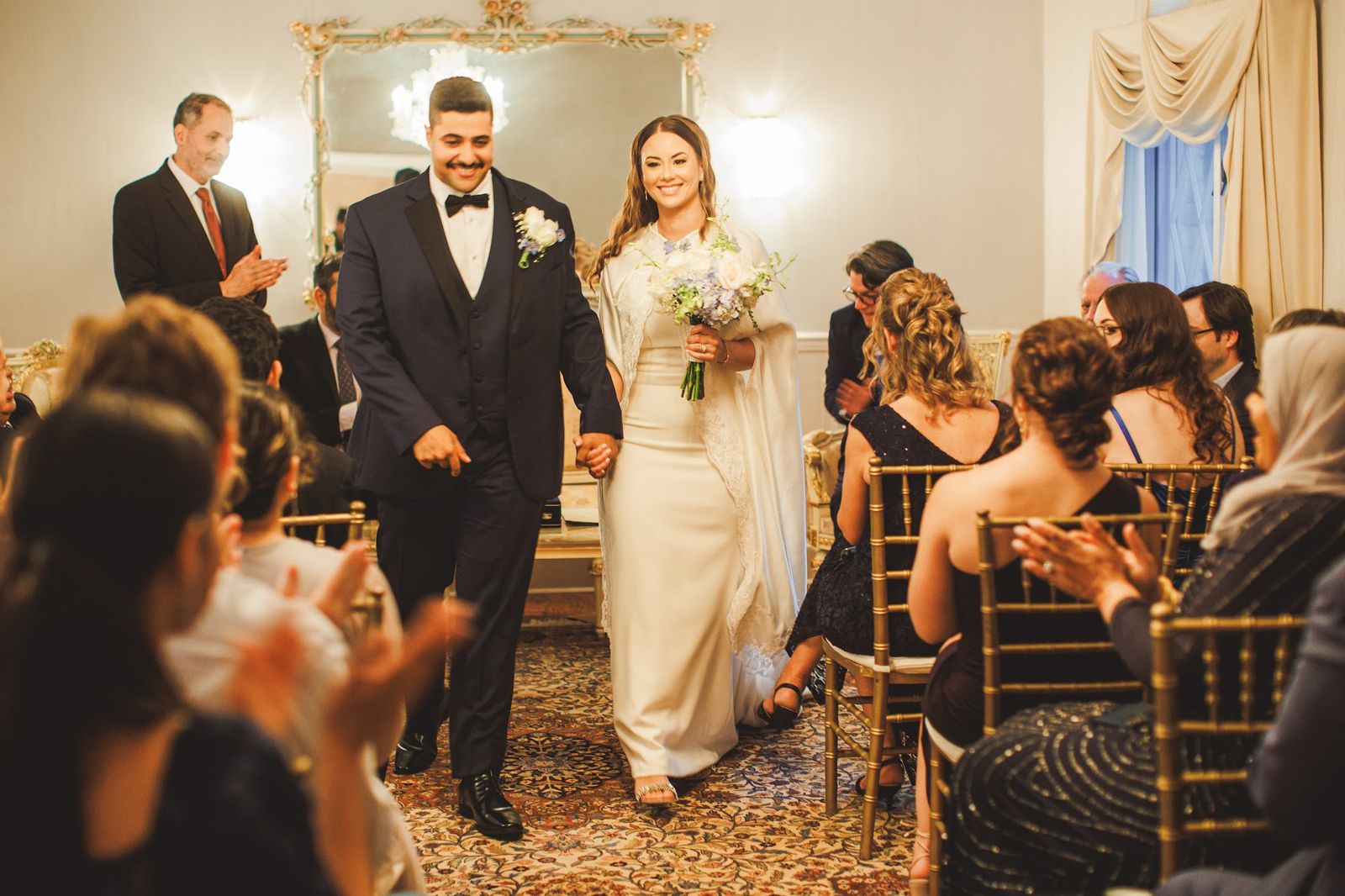 The couple walk down the aisle hand in hand while guests clap from their seats on both sides.