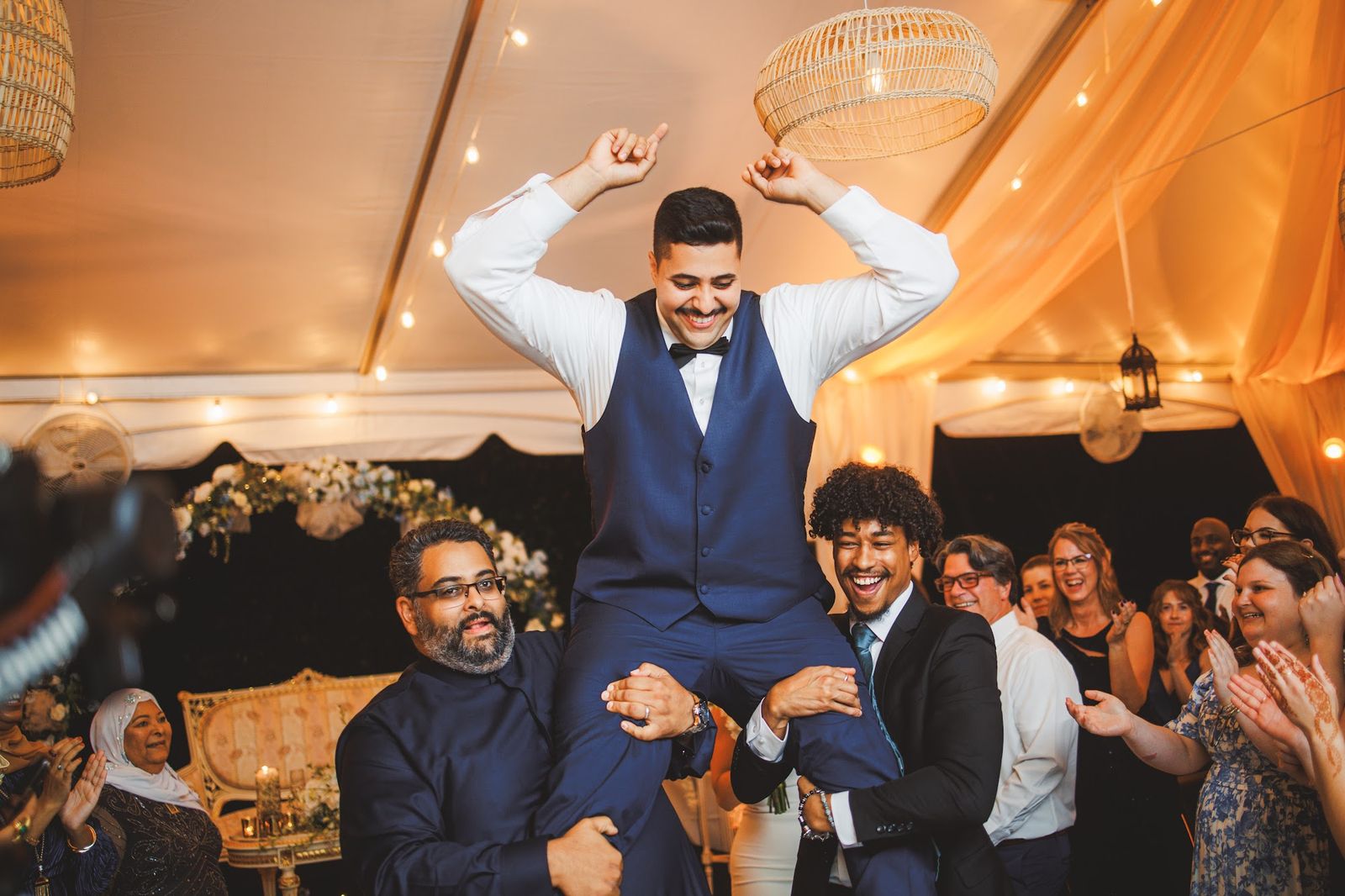 Two men carry the groom on their shoulders as he raises both arms and guests clap around them.