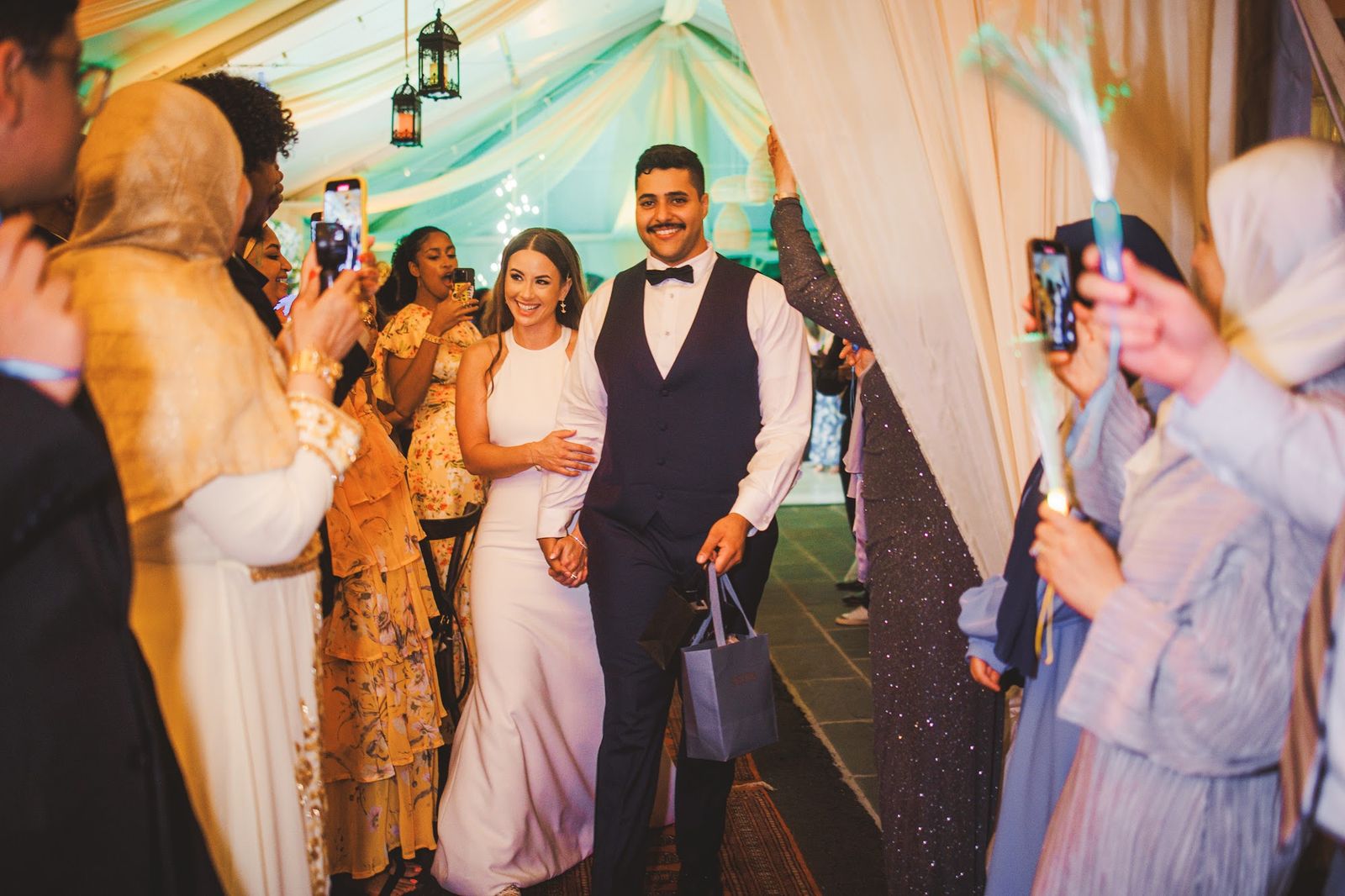 The couple walks arm in arm into the marquee between two rows of guests who film on their phones and hold glowing fiber-optic wands overhead.
