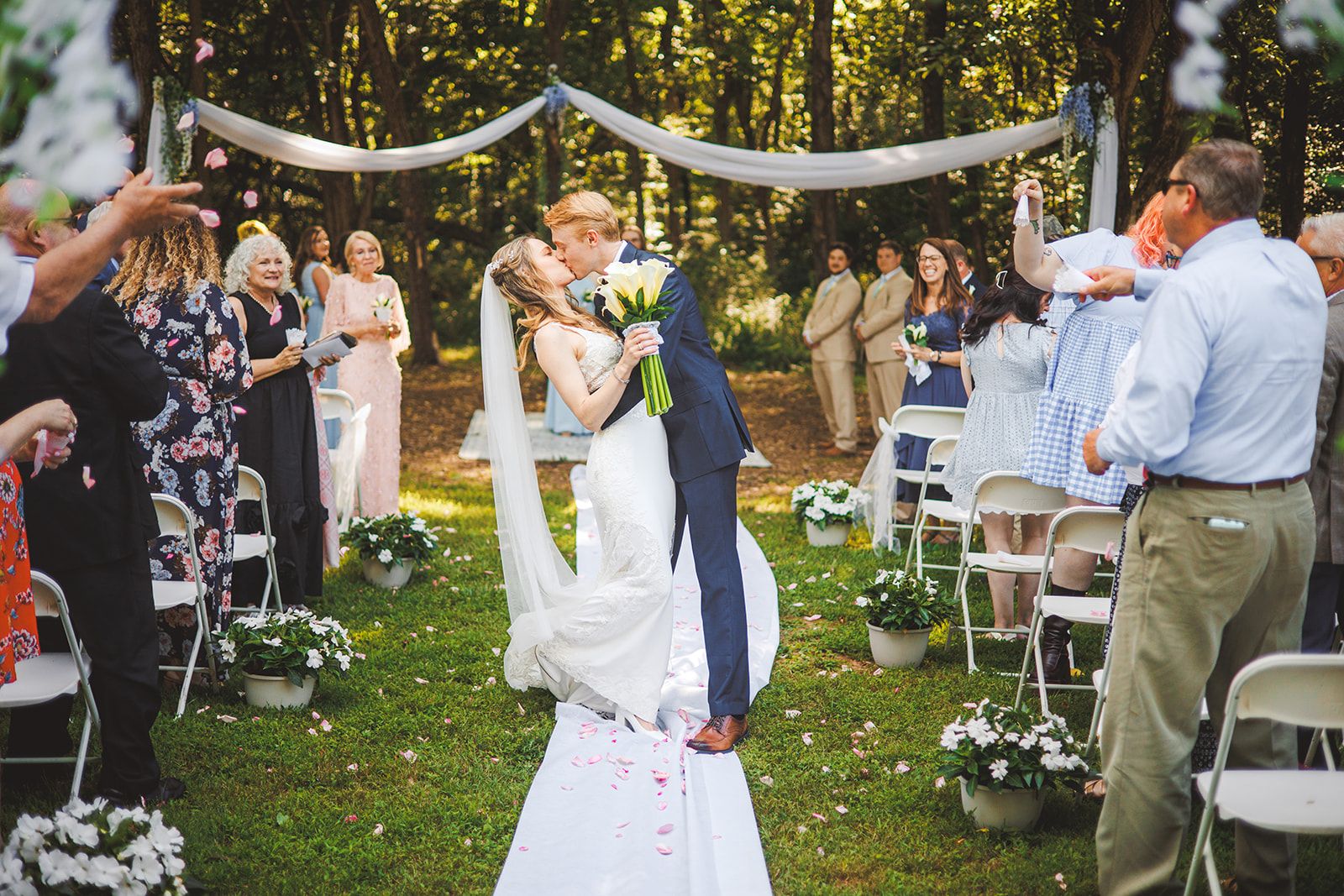 A couple kisses in the petal-strewn aisle under a draped arbor as a guest tosses pink petals into the air beside them.