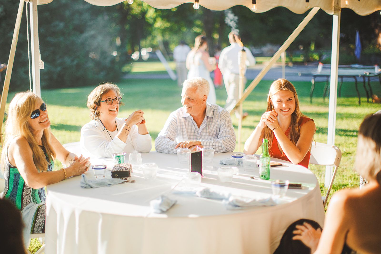 Guests sit around a round table under a tent in low golden light, one woman laughing with her hands raised as the man beside her turns to listen.