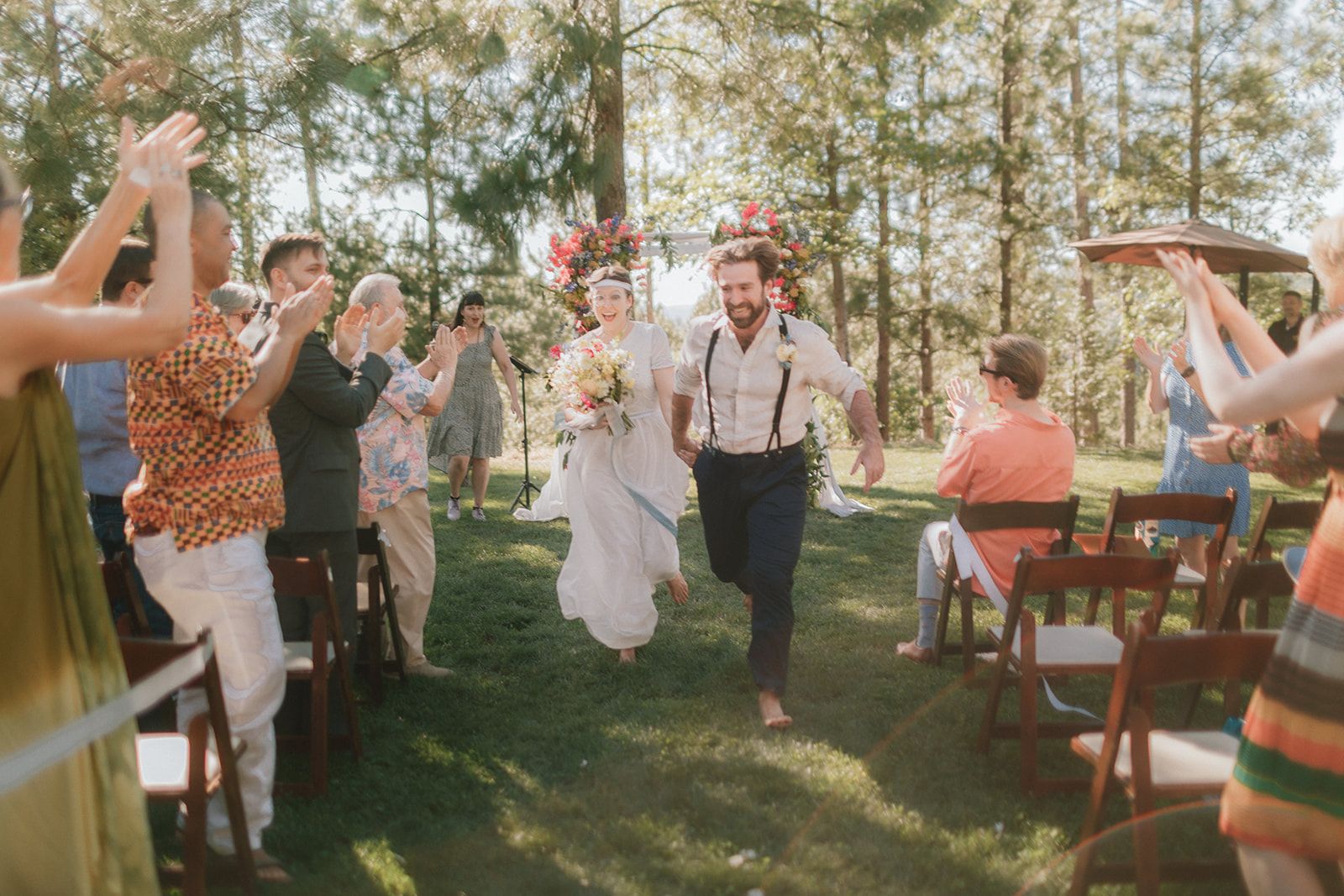 Guests clap with their hands raised on both sides of the aisle as the barefoot couple runs back down it, the bride in a flower crown holding her bouquet.