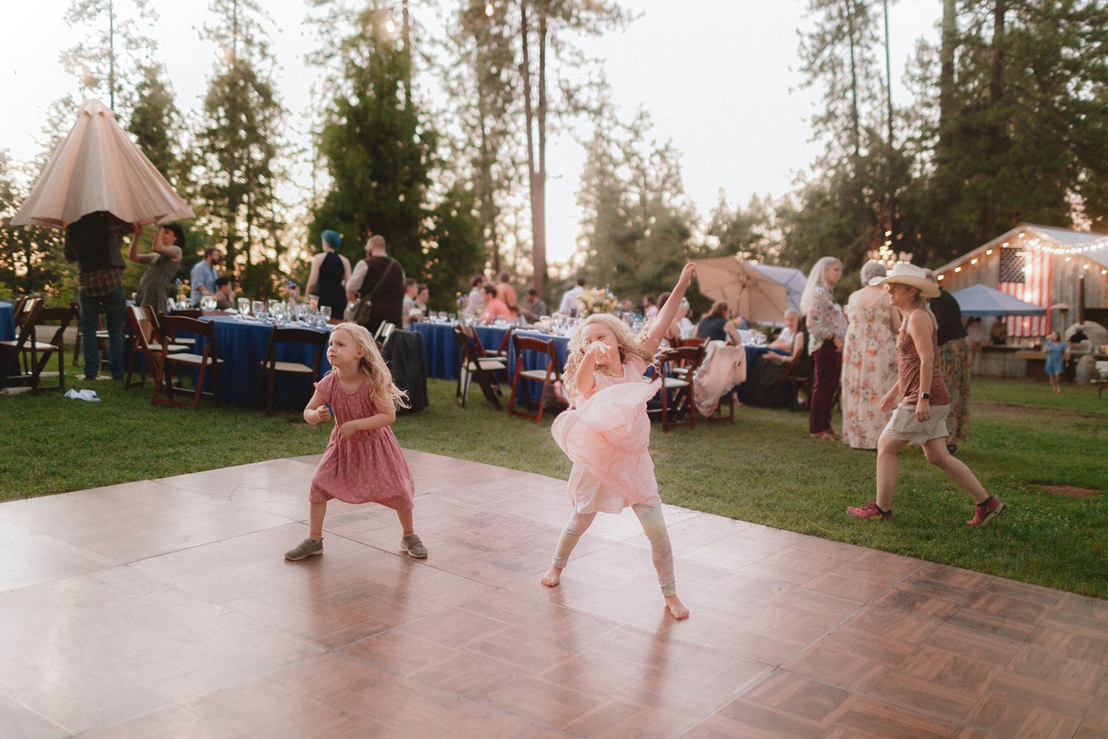 Two young girls dance on an outdoor wooden dance floor at dusk while guests sit at long tables behind them.