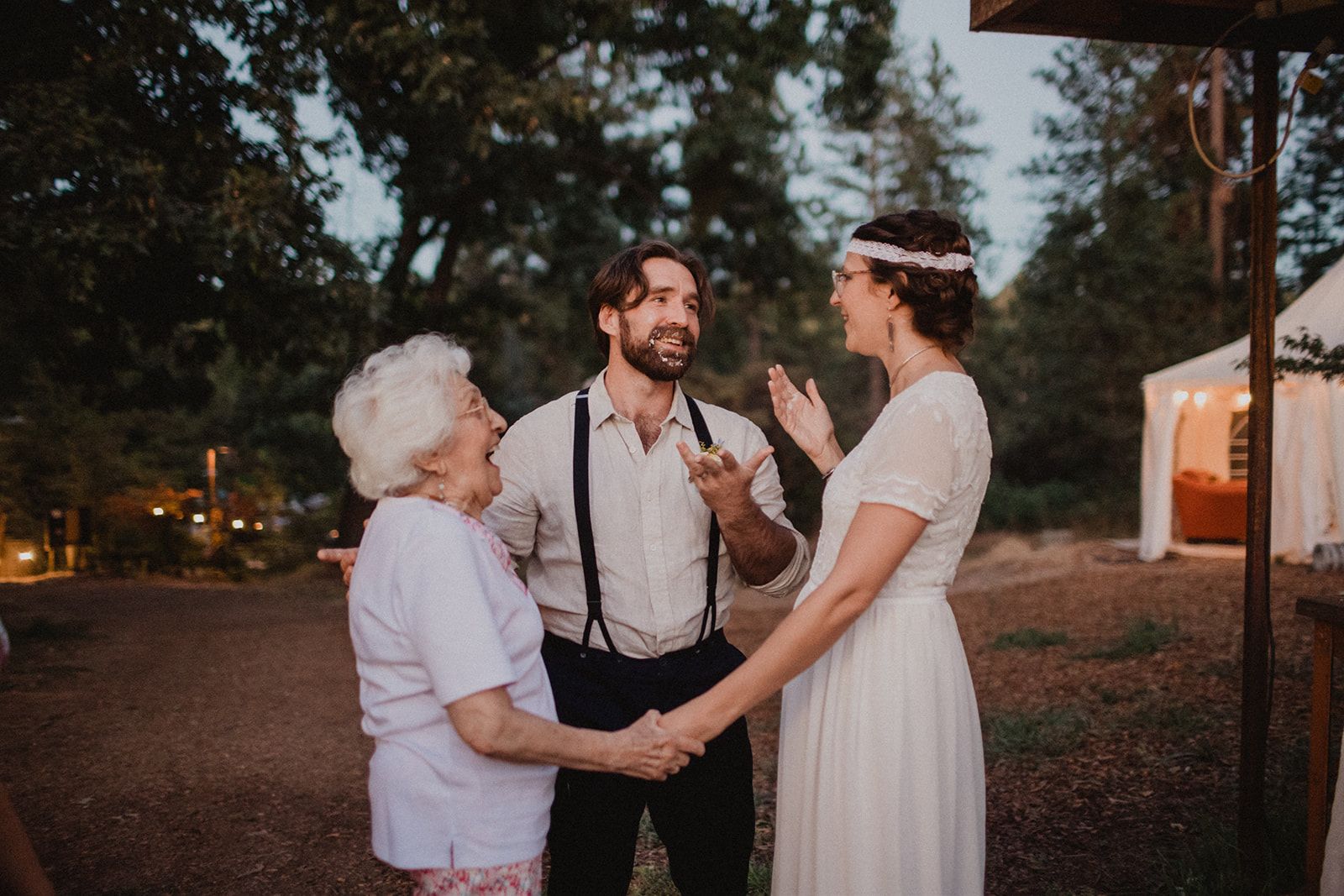 An older woman laughs face to face with the bride while holding her hand, and the groom stands between them with an arm around the older woman's back.