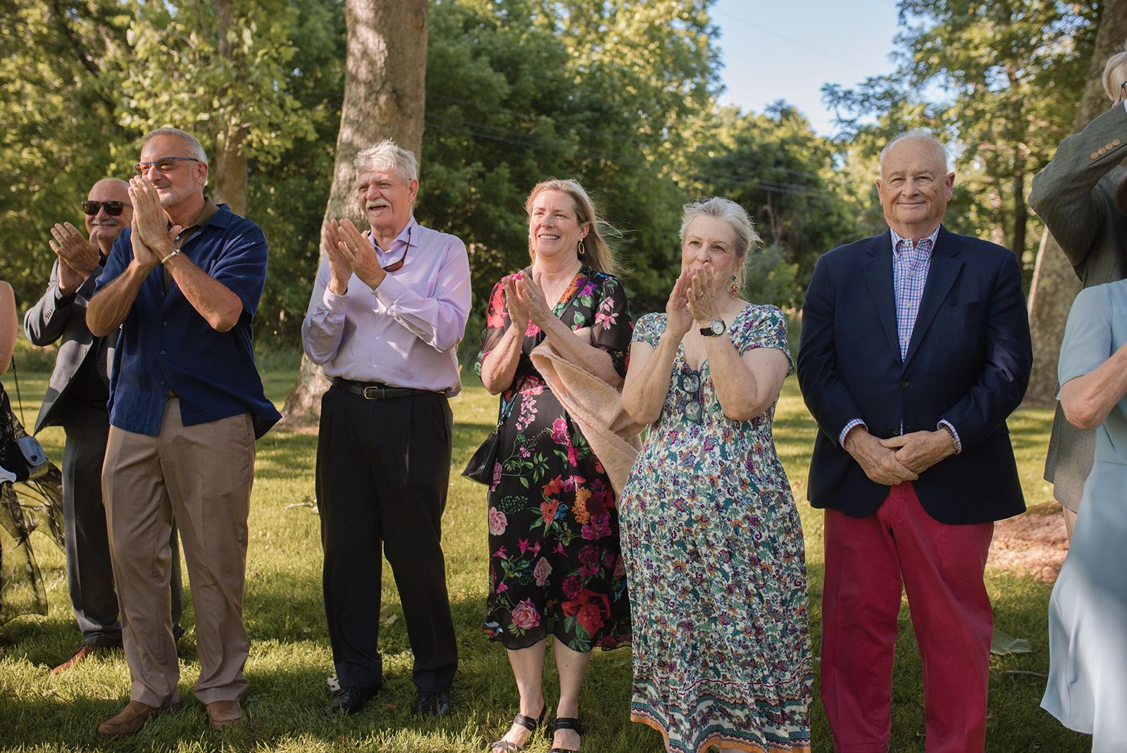 Wedding guests stand in a row on the grass, most of them applauding and smiling.