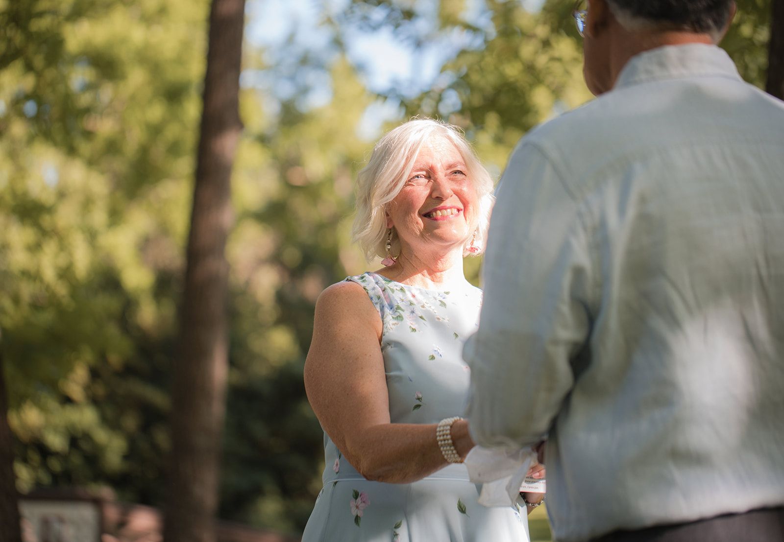 A woman with white hair in a pale blue dress smiles up at a grey-haired man who faces her with his back to the camera.