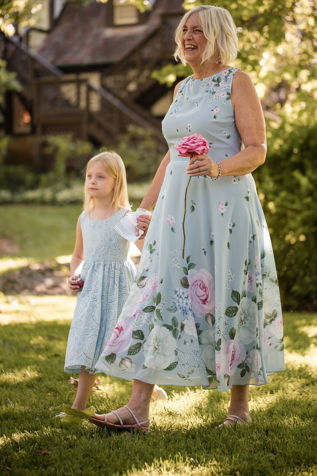A woman in a blue floral dress holds a pink rose and walks hand in hand with a young girl across the sunlit lawn.