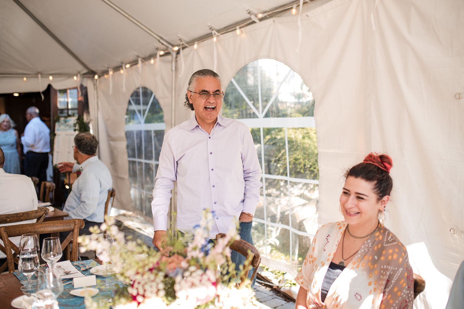 A man in a lavender shirt stands laughing behind a flower-covered table under a tent while a young woman at the right edge of the frame smiles.
