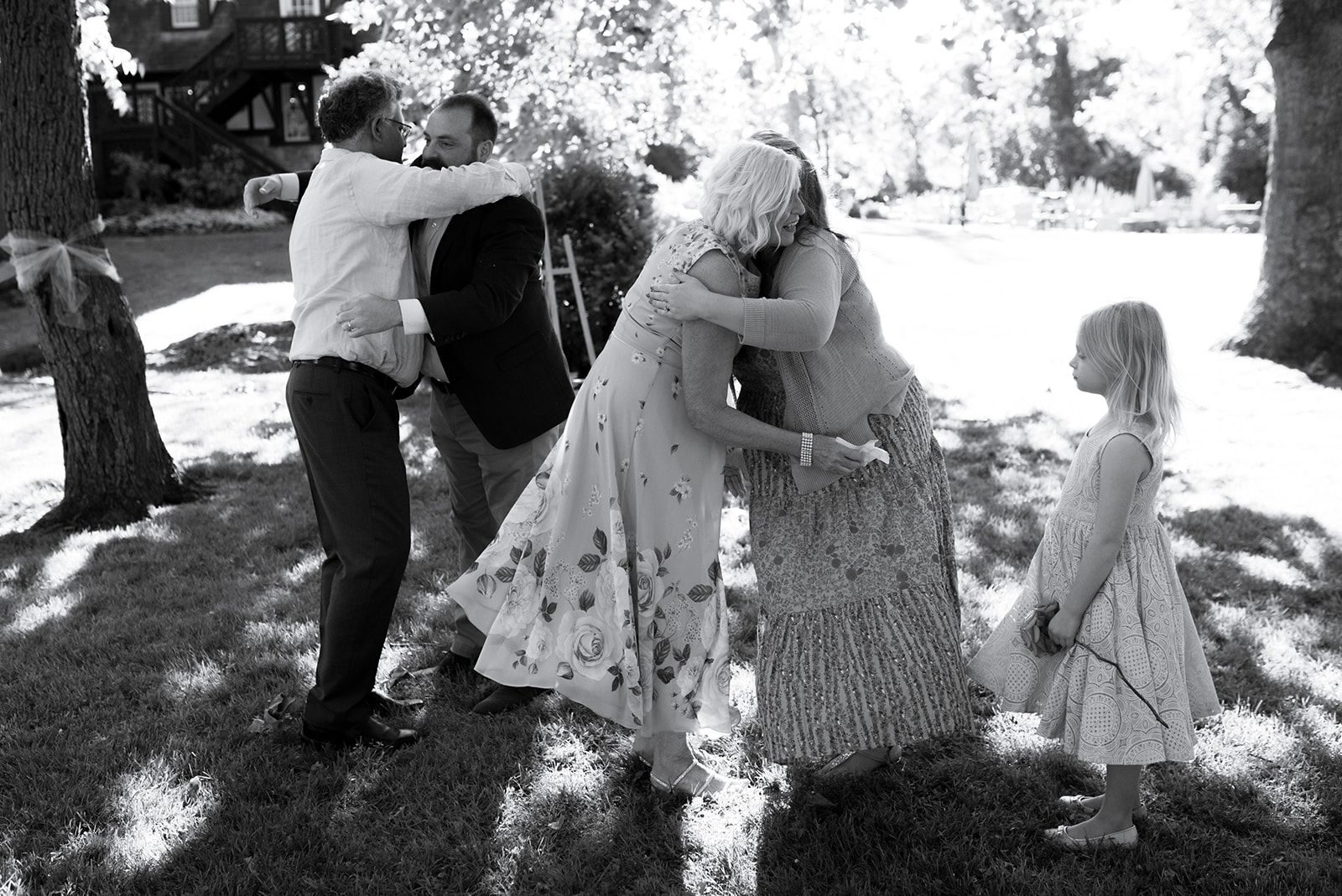 Two pairs of guests hug on a sunlit lawn while a young girl stands to the side watching them.