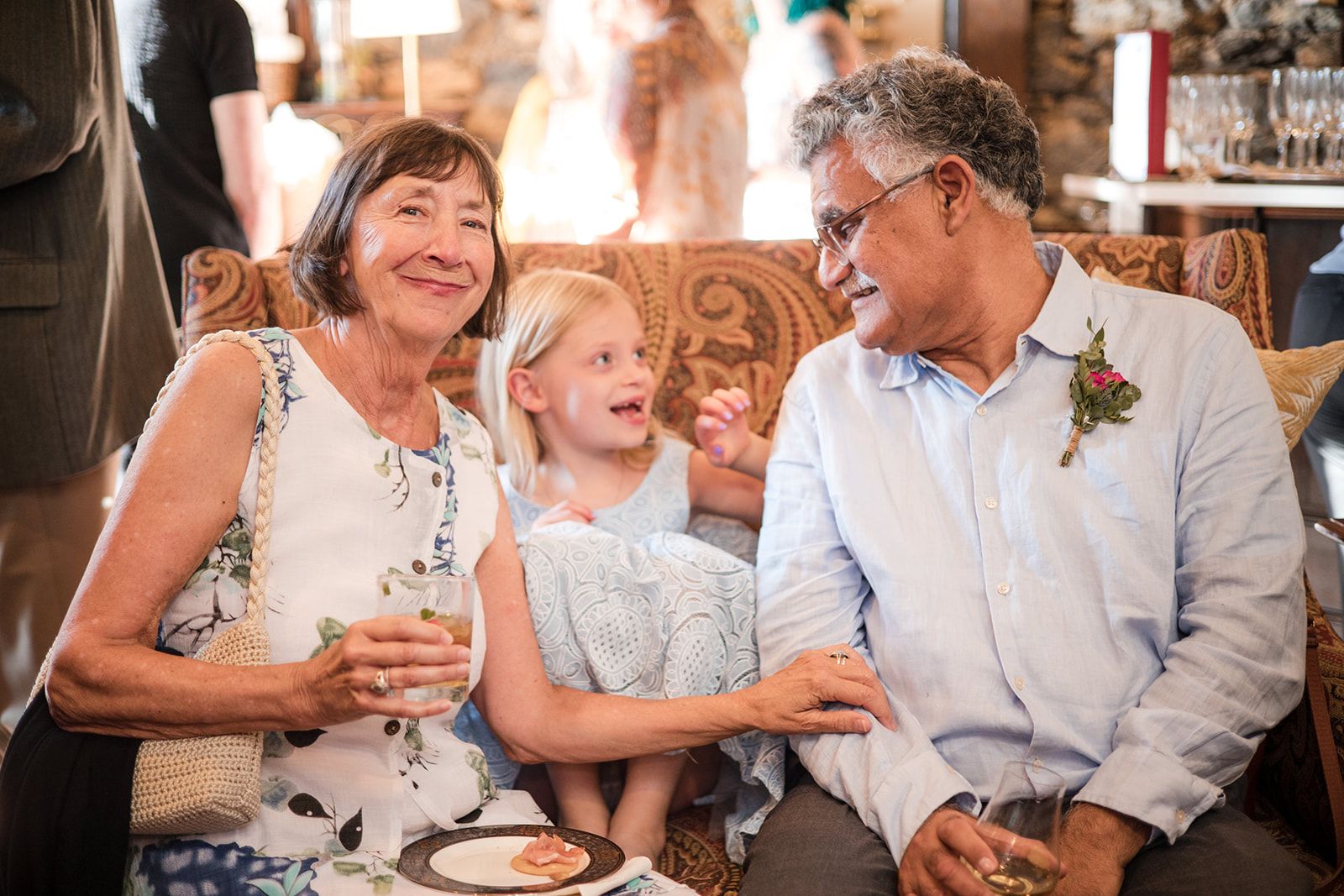 A little girl grins up at a gray-haired man laughing beside her on the couch while the woman on her other side smiles with a drink in hand.