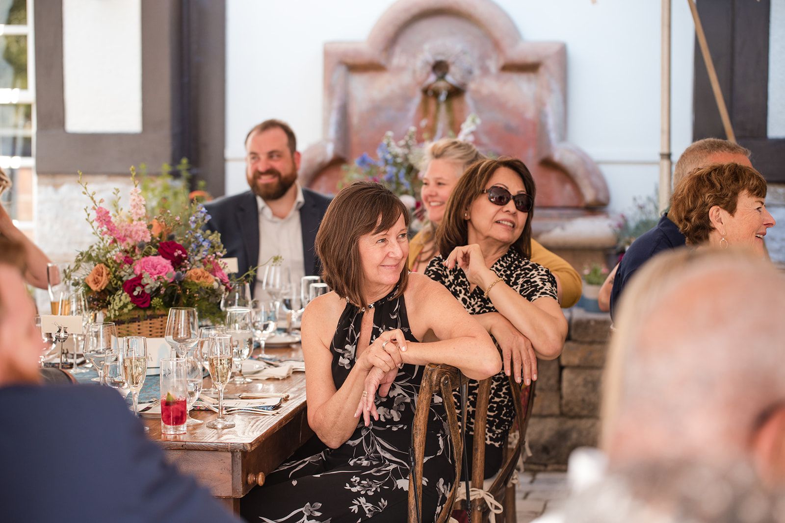 Two women turn in their chairs at a long dinner table, arms folded over the chair backs, watching something off frame.