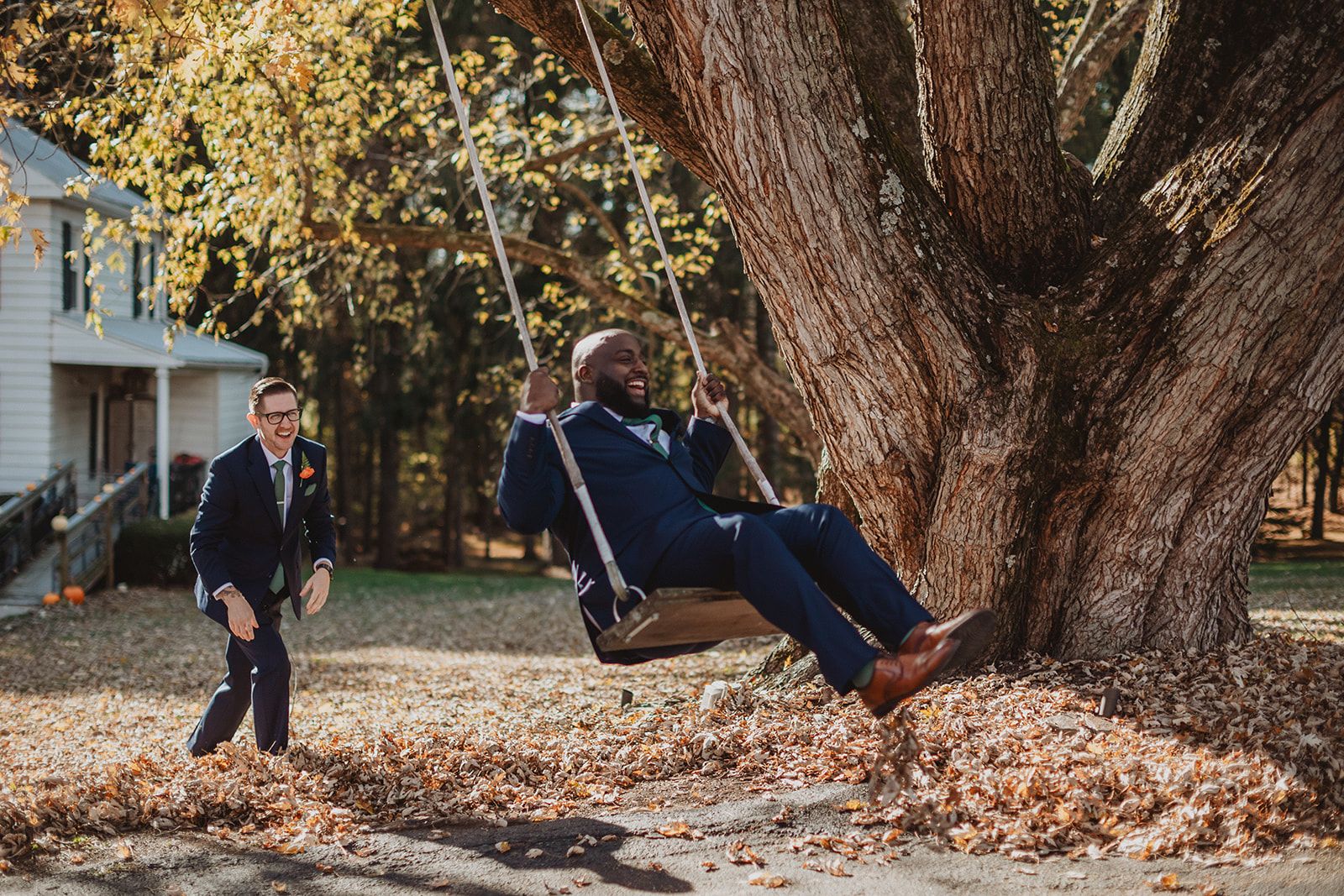 A man in a navy suit leans way back laughing on a rope swing hung from a massive tree while another man in a navy suit strides toward him through the fallen leaves.