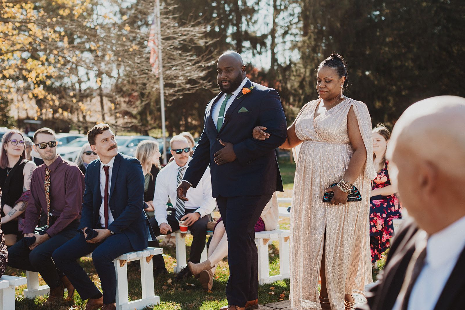 A man in a navy suit and green tie walks down the aisle arm in arm with a woman in a long champagne dress as seated guests watch from both sides.