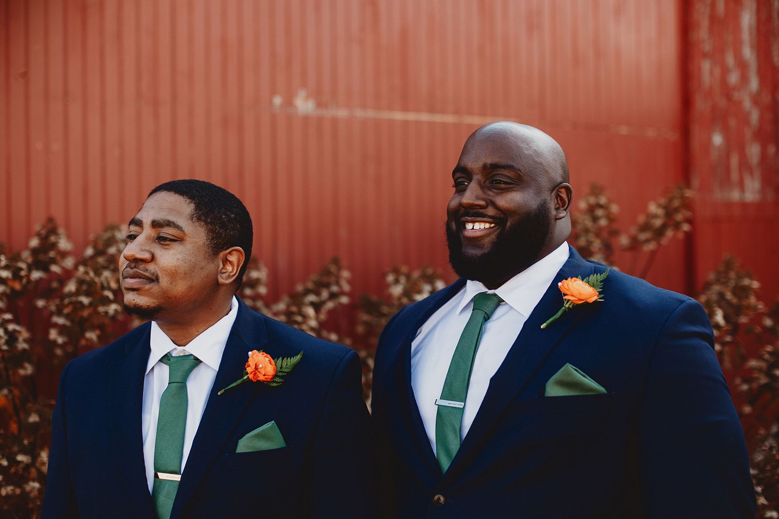 Two men in navy suits with green ties and orange boutonnieres stand side by side against a red corrugated wall, the one on the right grinning wide at something off camera.