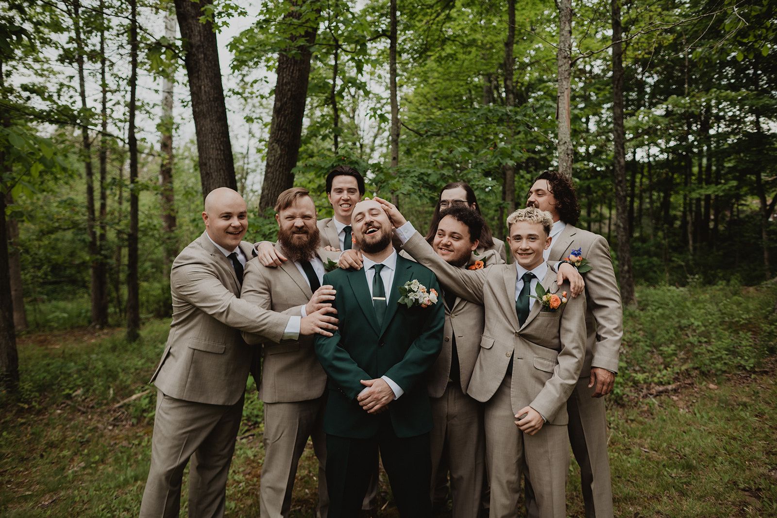 A wedding party in matching tan suits piles in around the groom in a dark green suit, laughing while one of them lays a hand on top of his bald head.