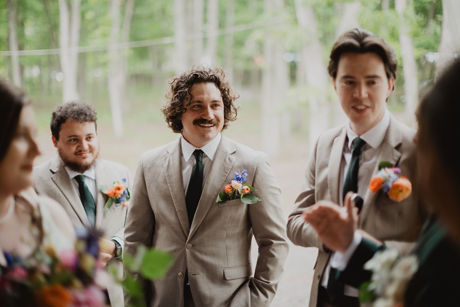 A man in a tan suit grins between two other men in matching tan suits, with out of focus guests and bouquets framing the foreground.