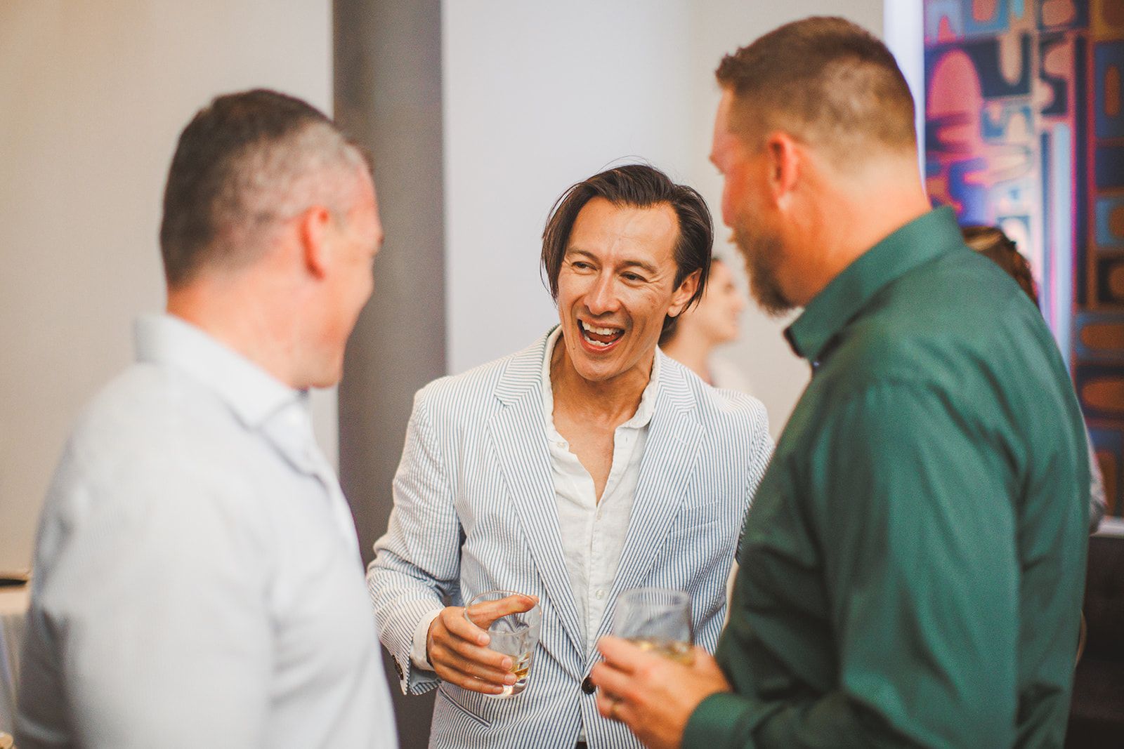 Three men stand talking with drinks in hand while the one facing the camera laughs mid conversation.