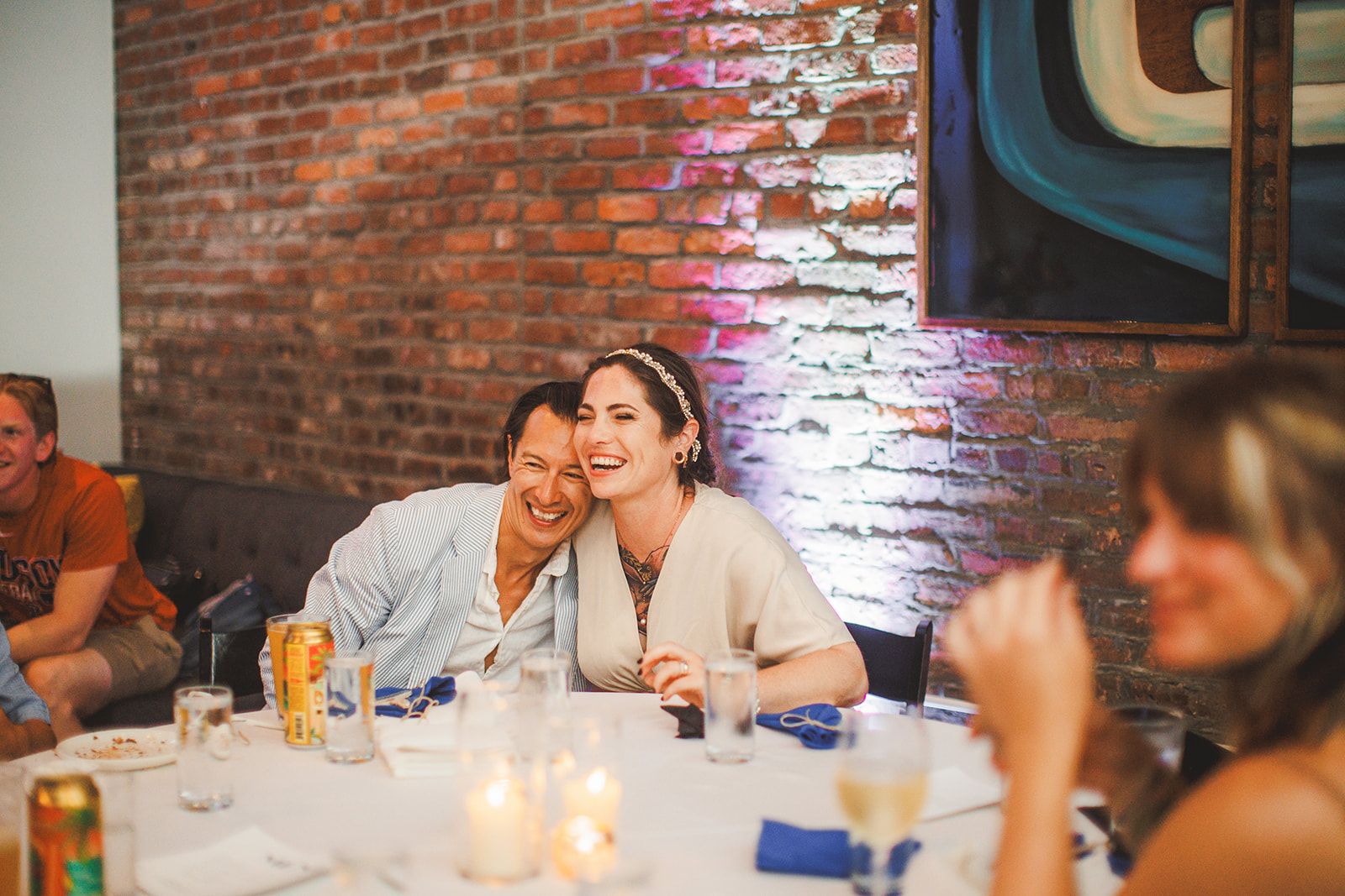 A man presses his cheek to the bride's as she laughs at a reception table.