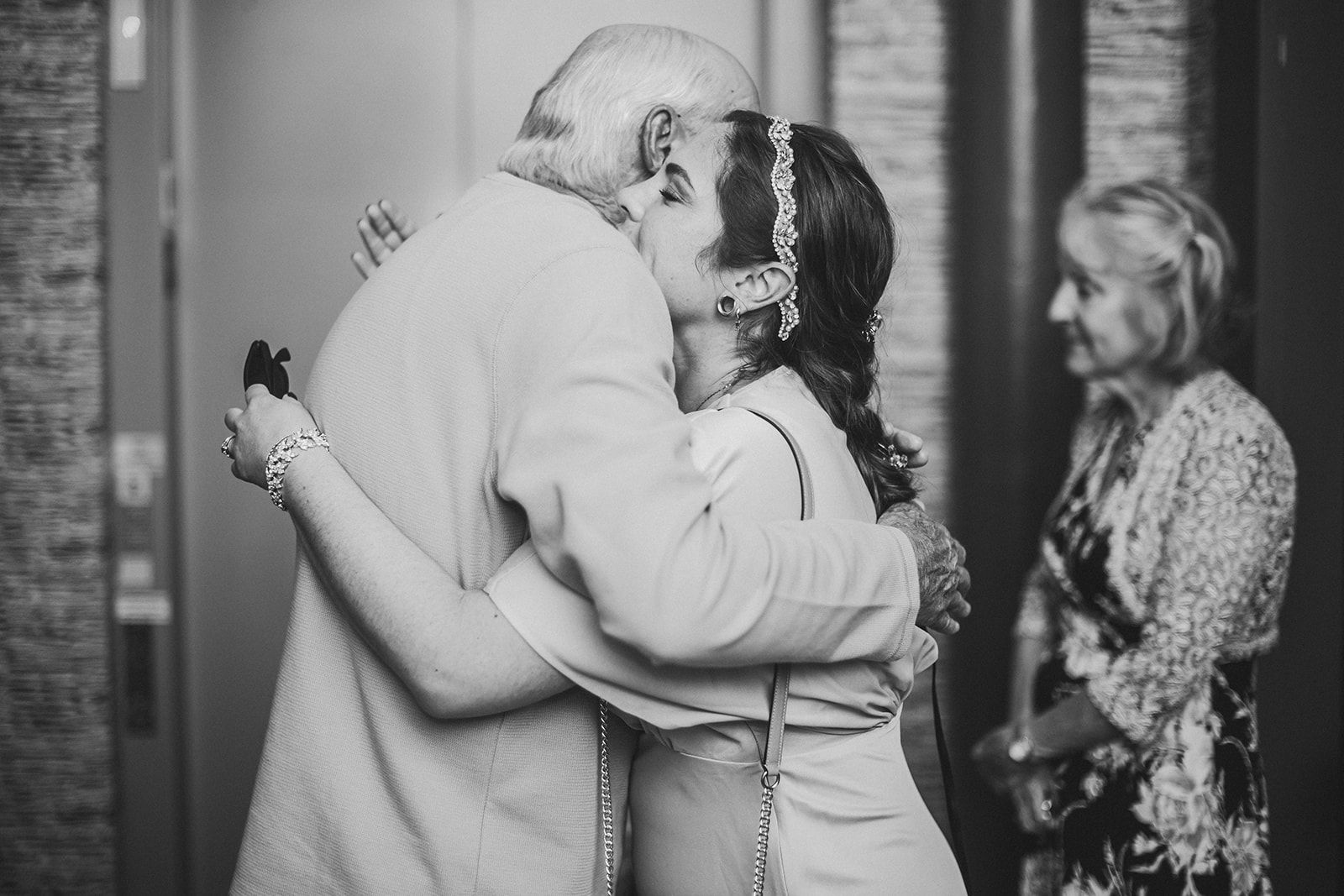 The bride wraps a white haired older guest in a tight hug while another woman watches from behind them.