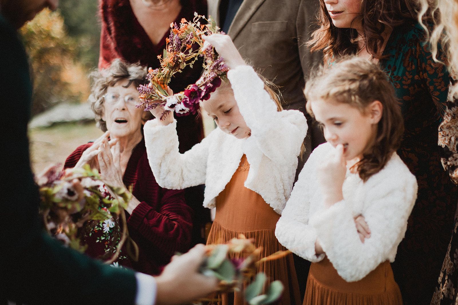 A girl raises a flower crown above her head while another girl holds a hand to her mouth and a woman behind them reacts with her mouth open.