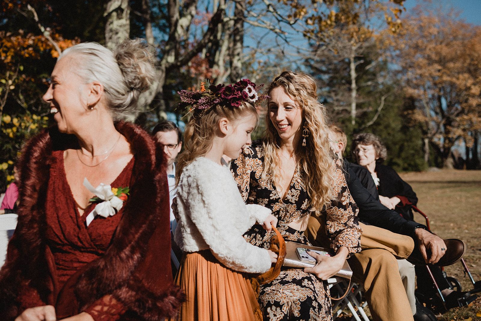 A woman with white hair in a burgundy dress laughs while a girl in a flower crown stands beside a curly-haired woman who smiles down at her.