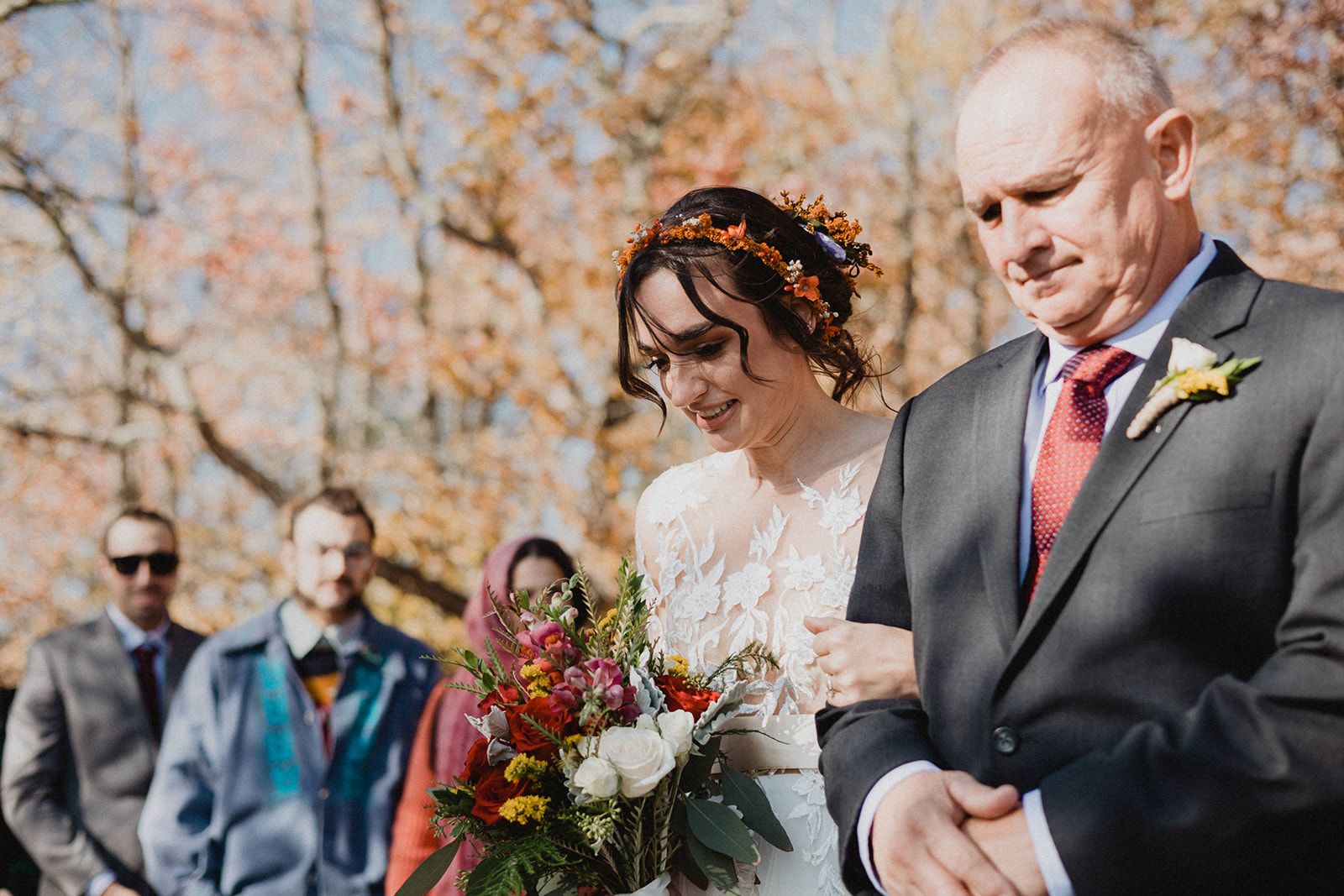 A bride holds a grey-suited man's arm and looks down with a small smile as guests stand out of focus behind them.