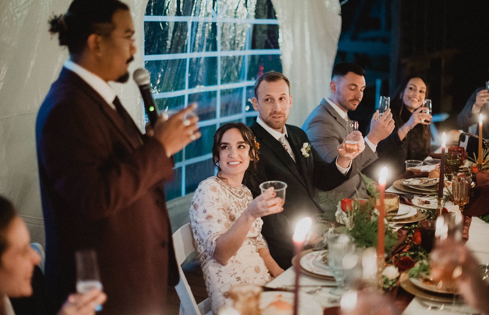 A man in a dark suit speaks into a microphone beside the candlelit head table while the seated bride raises her cup and guests down the table lift their glasses toward him.