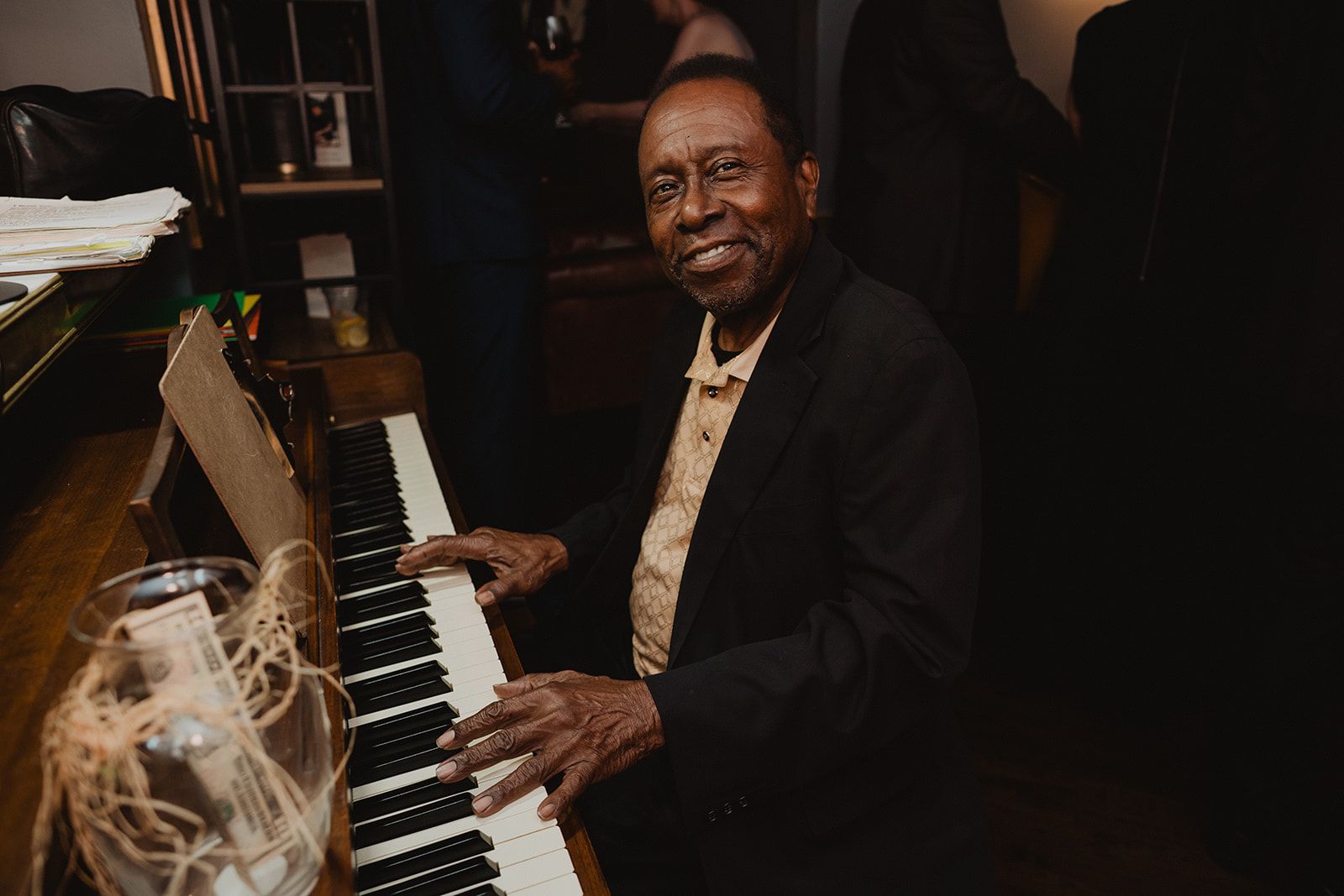 An older man in a black suit jacket sits at an upright piano with both hands on the keys and smiles toward the camera.