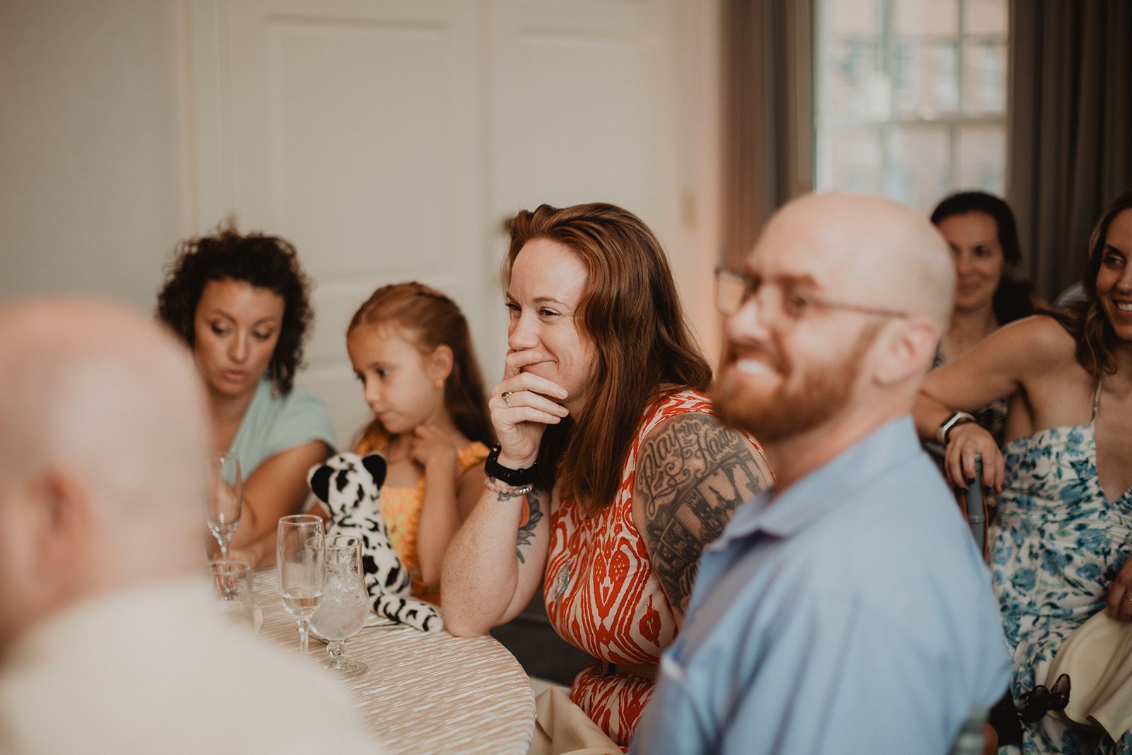 A woman at the long table presses a hand over her mouth watching something off camera while a young girl beside her holds a stuffed animal.