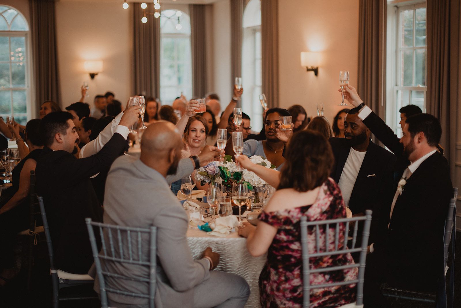 Guests packed around a long banquet table lift their glasses together during a toast.