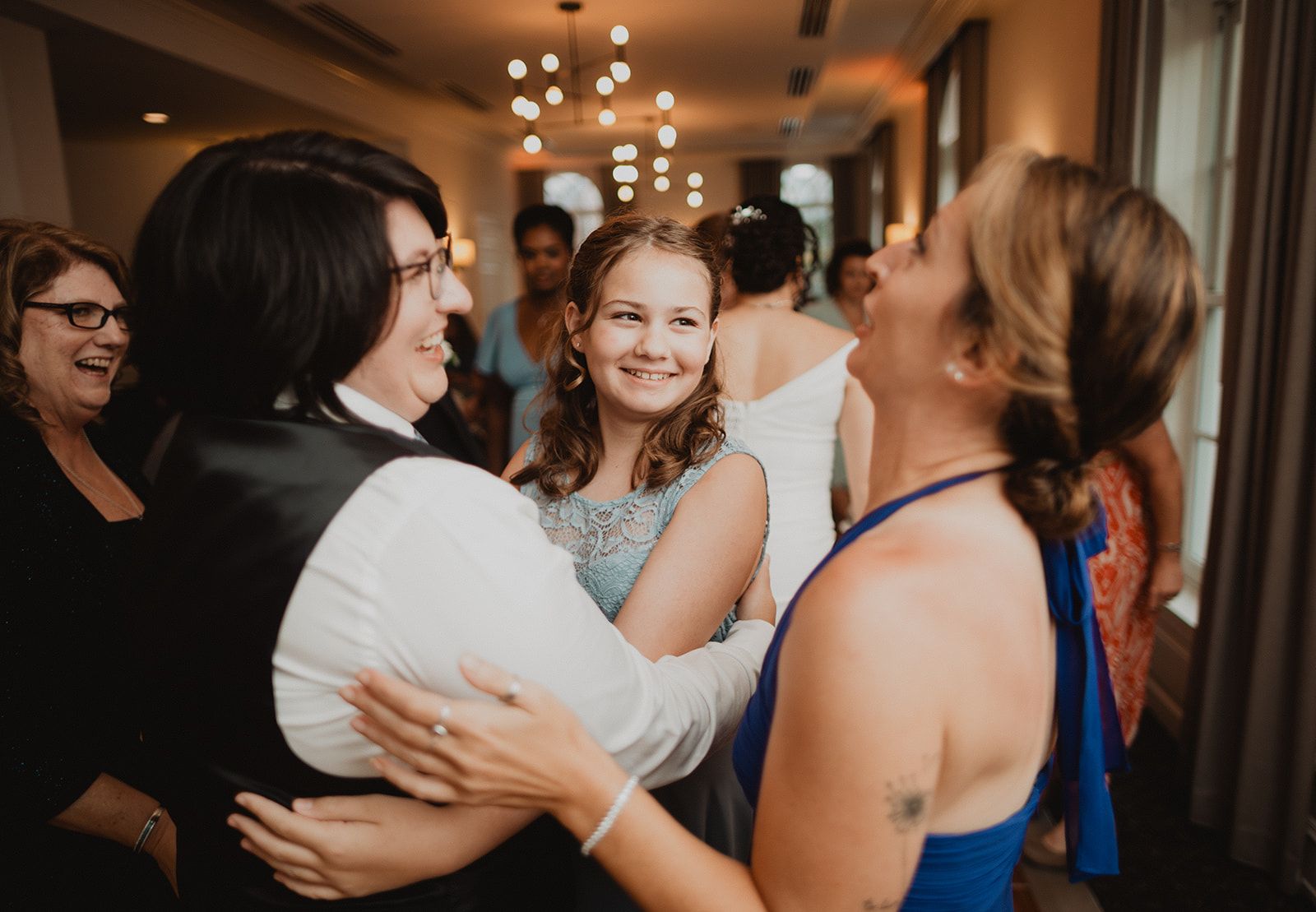Guests fold each other into a hug at the reception while a girl in the middle smiles and a woman beside them laughs with her head tipped back.