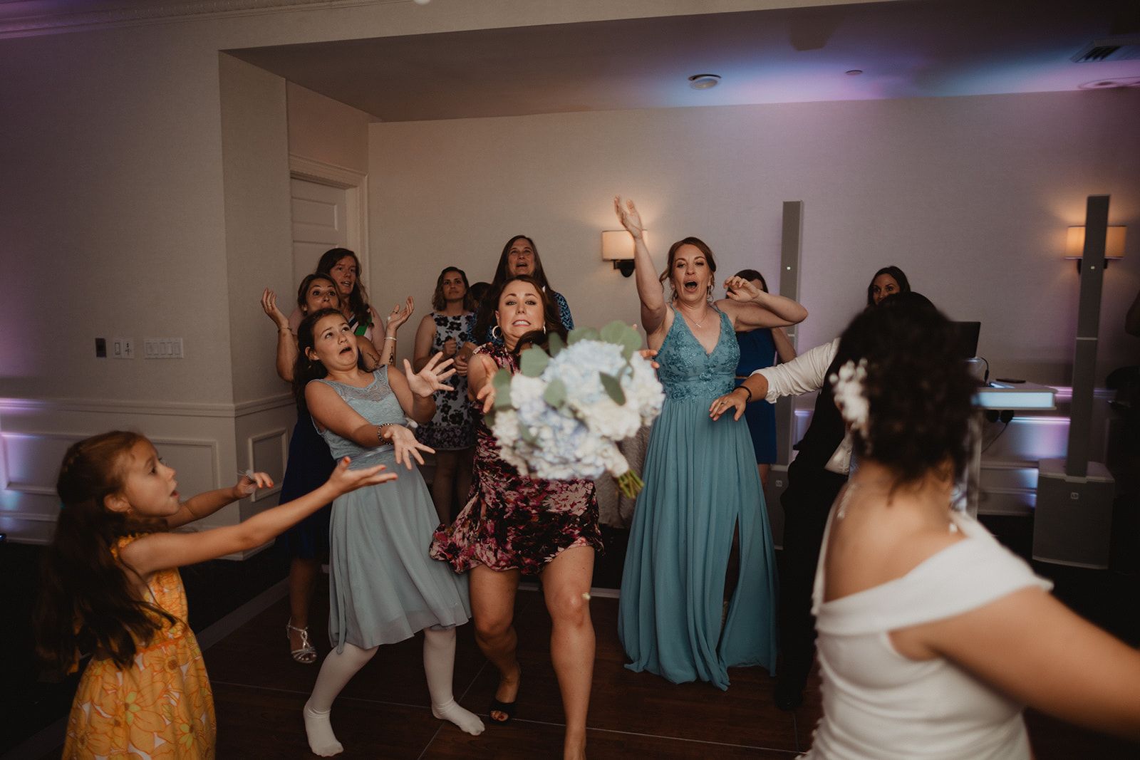 A young girl in a yellow dress and a cluster of women lunge with open hands as the tossed bouquet drops among them.