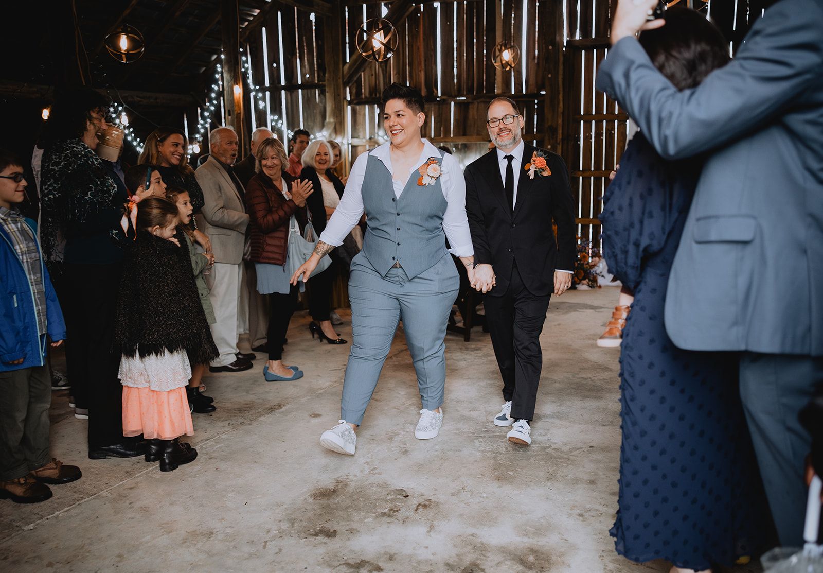 Two people in wedding clothes stride side by side through a barn while the guests lining the way clap and a small girl in a peach skirt watches from the front.