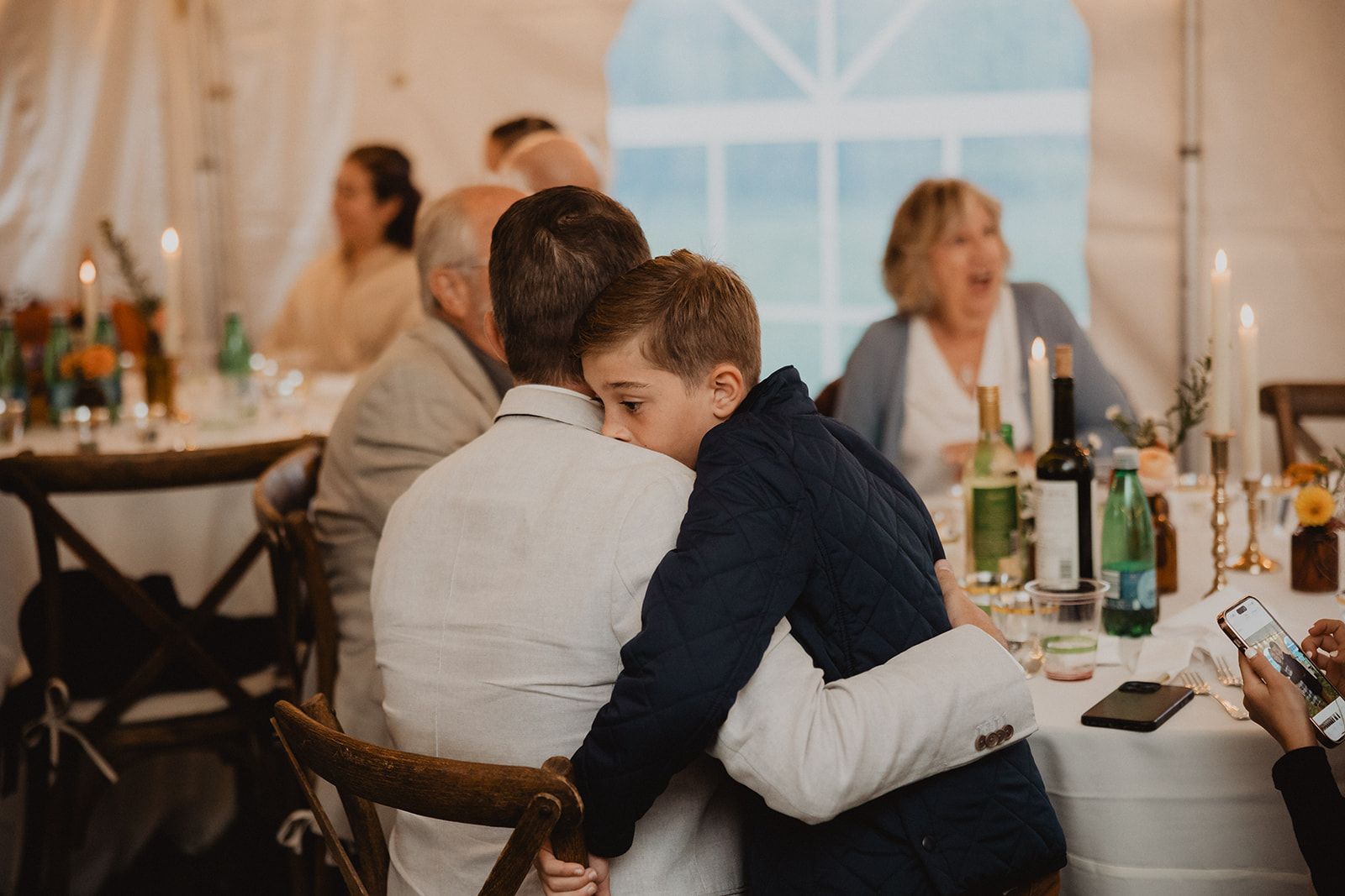 A boy in navy leans into a seated man in a white shirt at the reception table with his cheek against the man's back and an arm wrapped around him, while a woman behind them laughs.